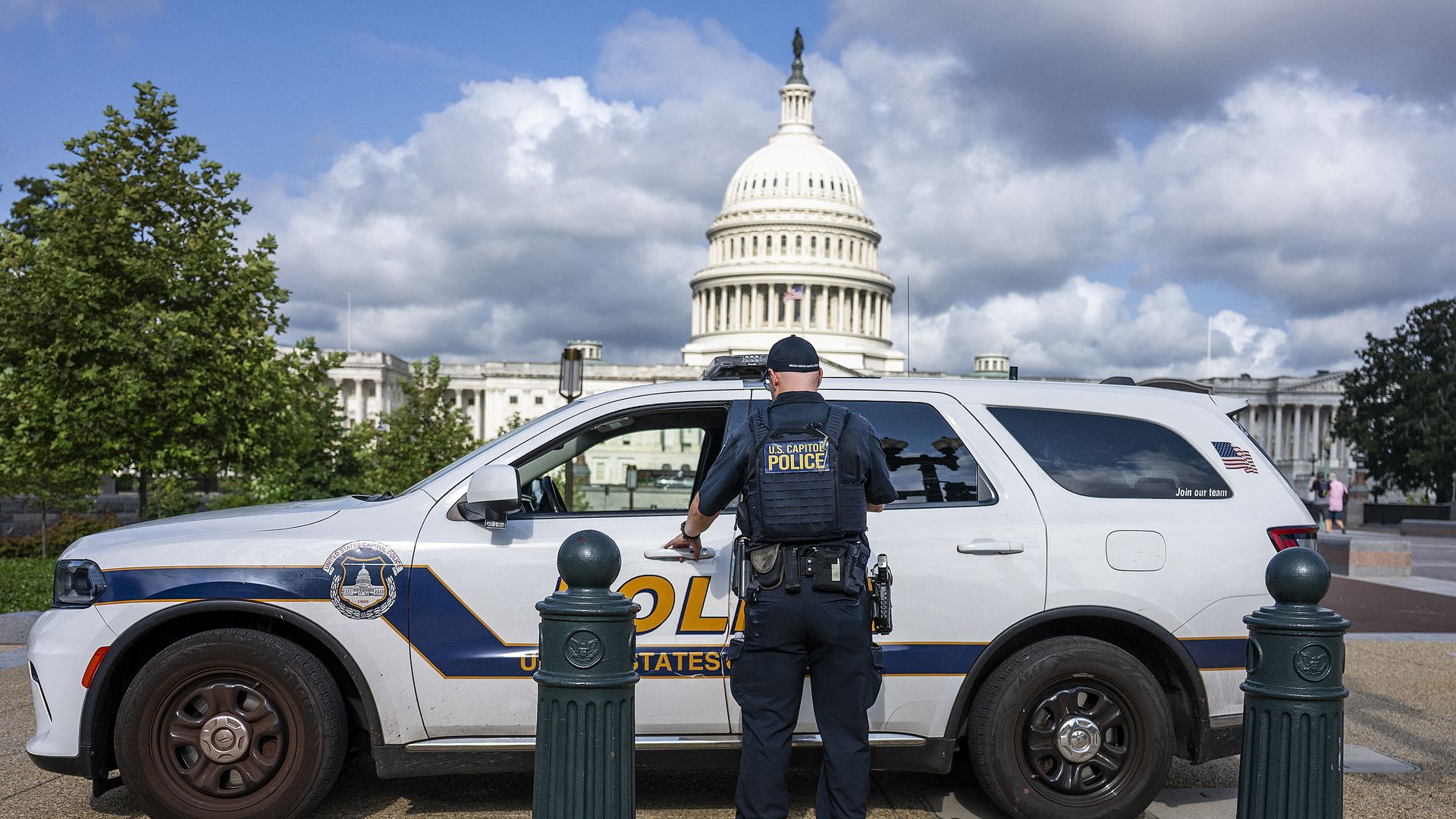 U.S. Capitol Police officer in dark uniform opening white police SUV with U.S. Capitol building and partly cloudy sky in background.