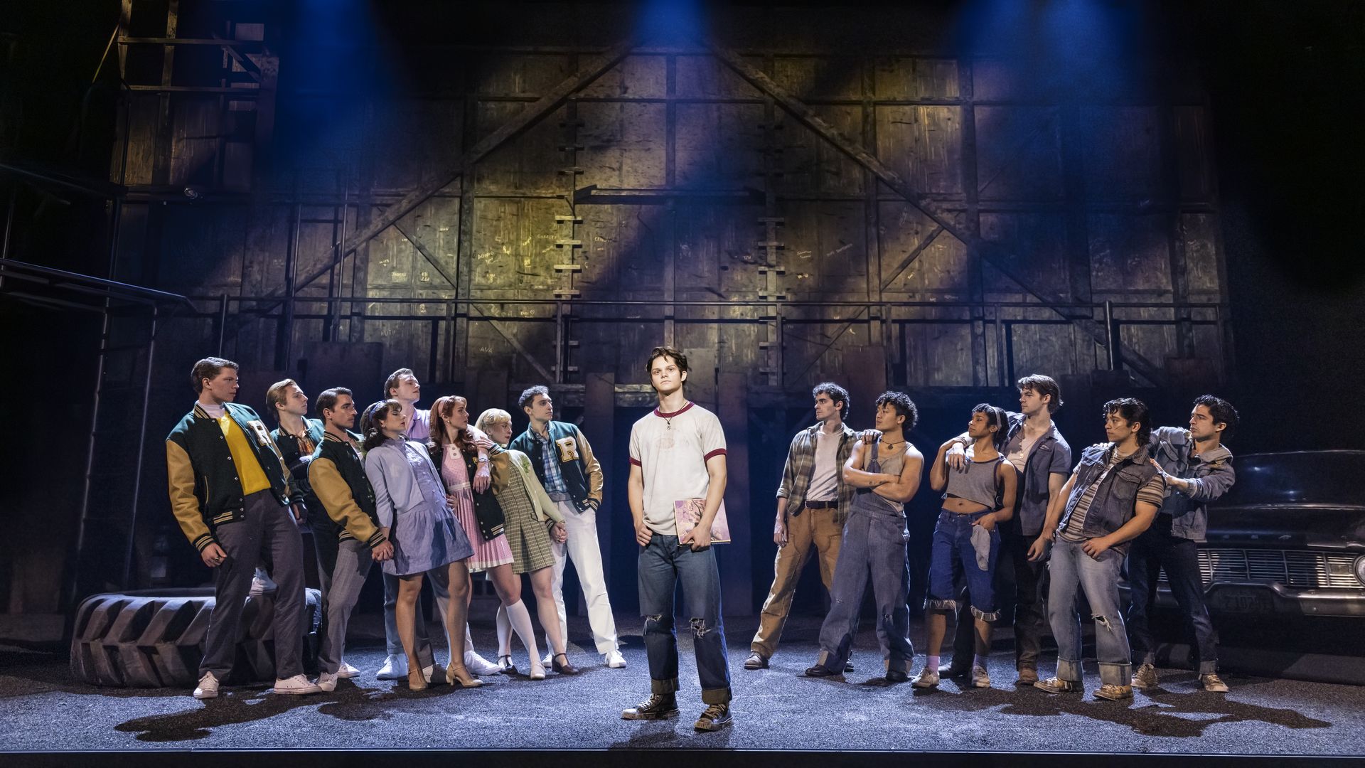 A group of 14 young actors stand on stage under blue spotlights. One stands center holding a book, others face him. They wear casual 50s-style clothes, with a dark industrial backdrop.