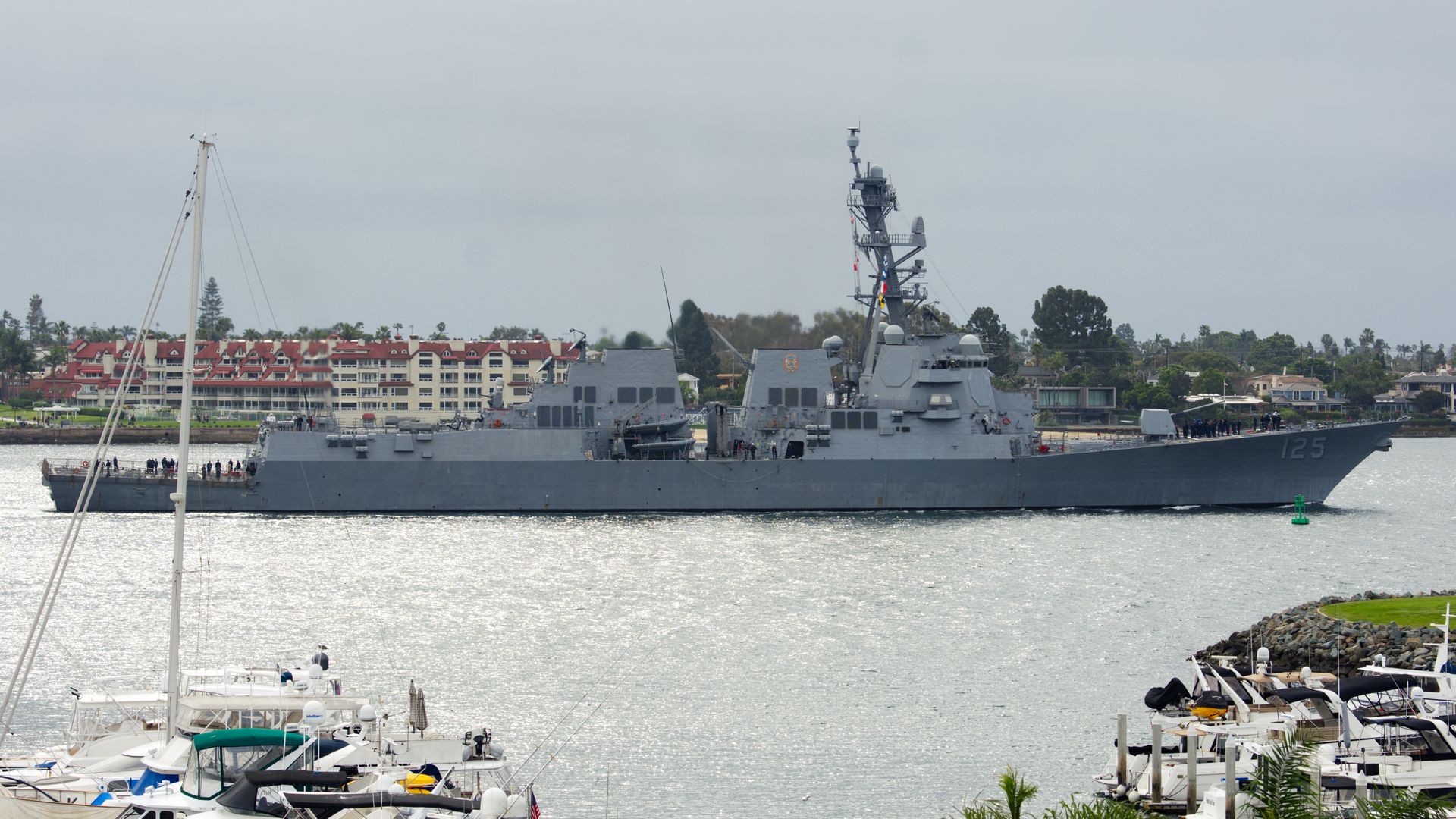 Gray naval warship marked 125 sails near a marina filled with white boats, with a cloudy sky and buildings with red roofs in the background.