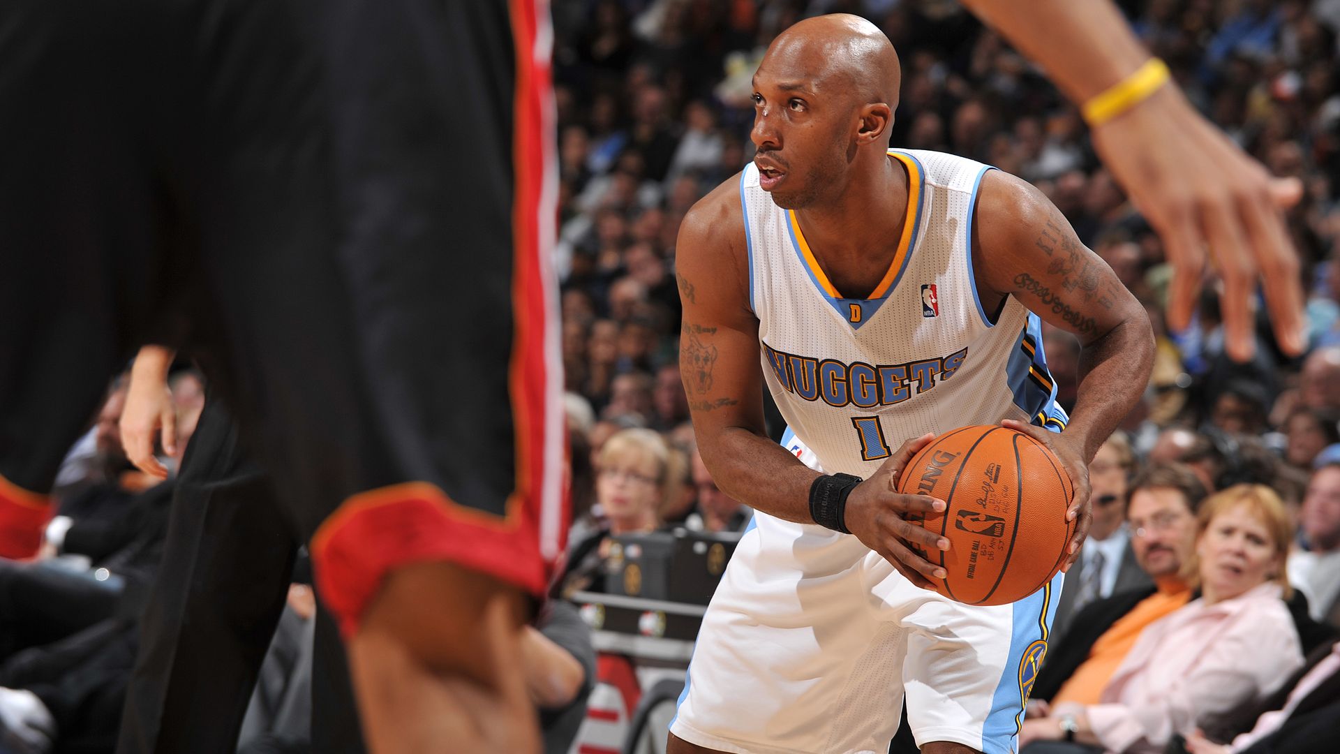A man in a white jersey with baby blue trimming on its side holds an orange basketball, preparing to pass it, while inside a large arena. A crowd can be seen in the background.