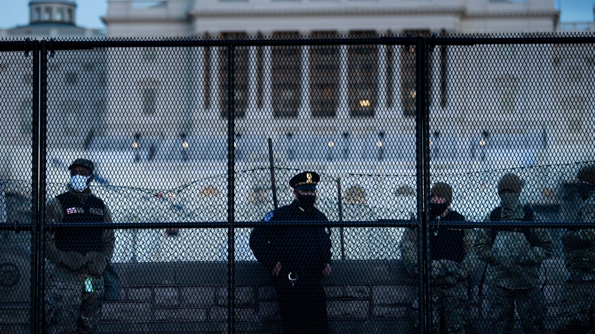 Photo of four law enforcement officers in uniform standing behind a fence that surrounds the U.S. Capitol