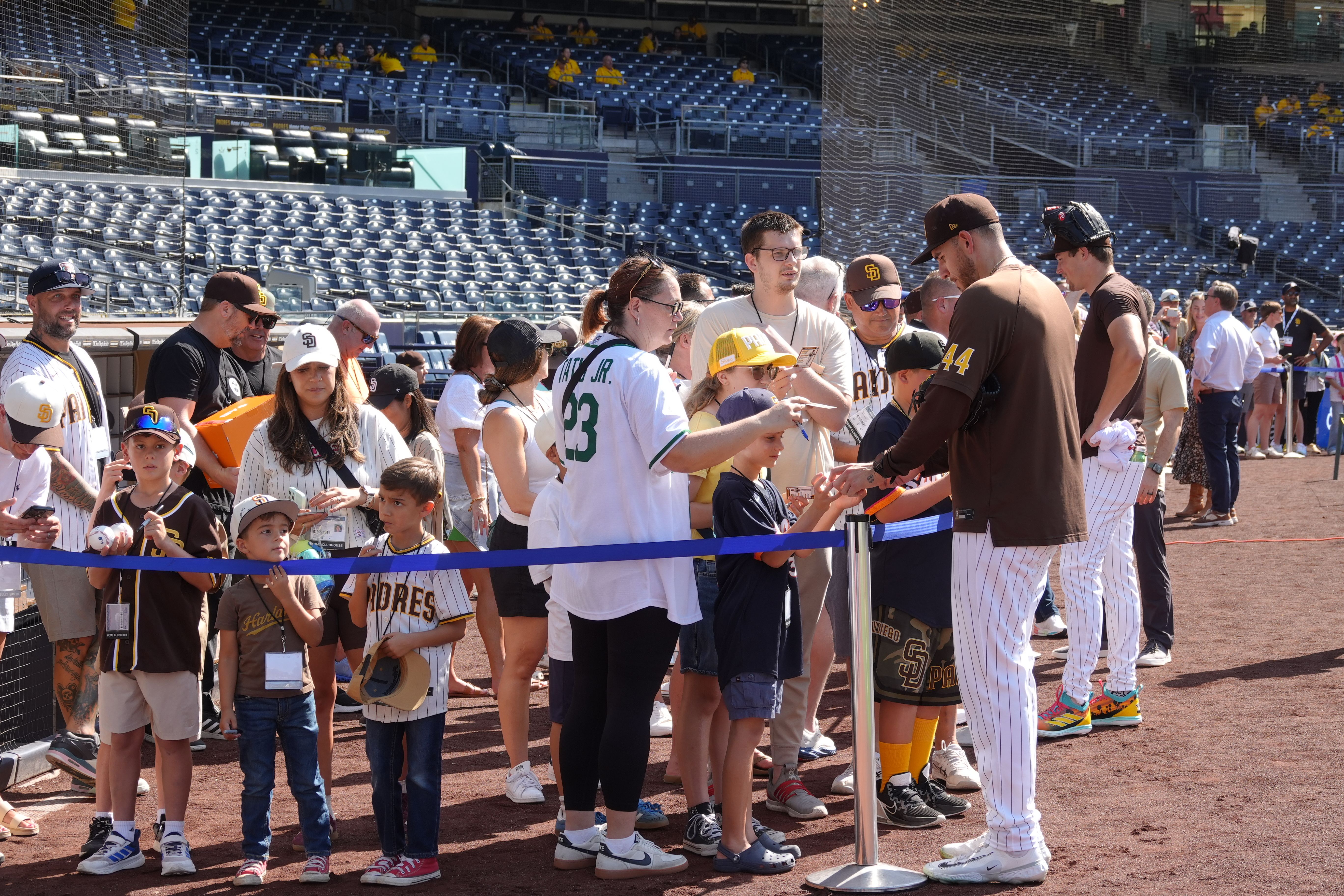 Fans, mostly kids, line up for Padres autographs along a rope barrier at a sunny baseball stadium; players in brown and white uniforms, blue seats and netting in the background.