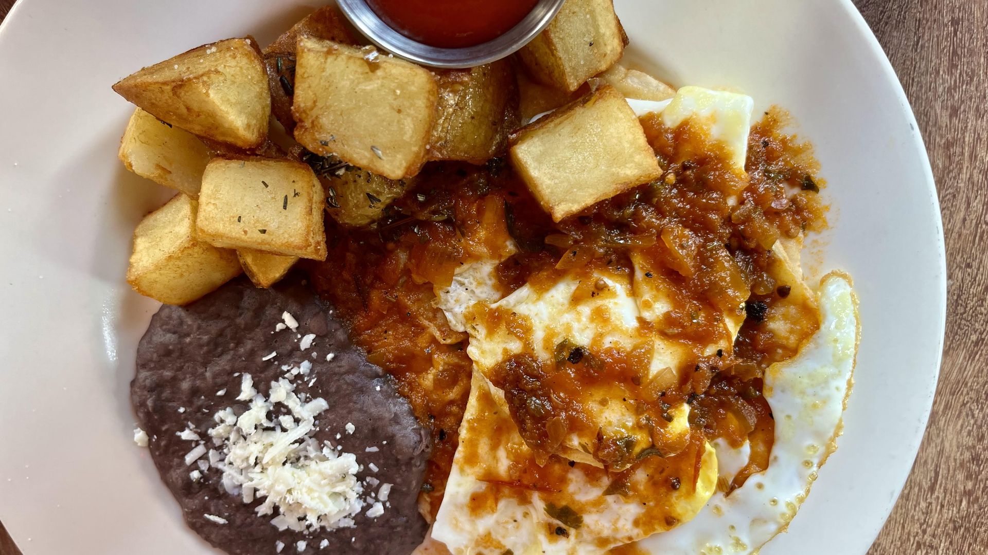 A white plate with fried potatoes, refried black beans topped with white cheese, and eggs covered in red salsa, with a small cup of ketchup on the side.