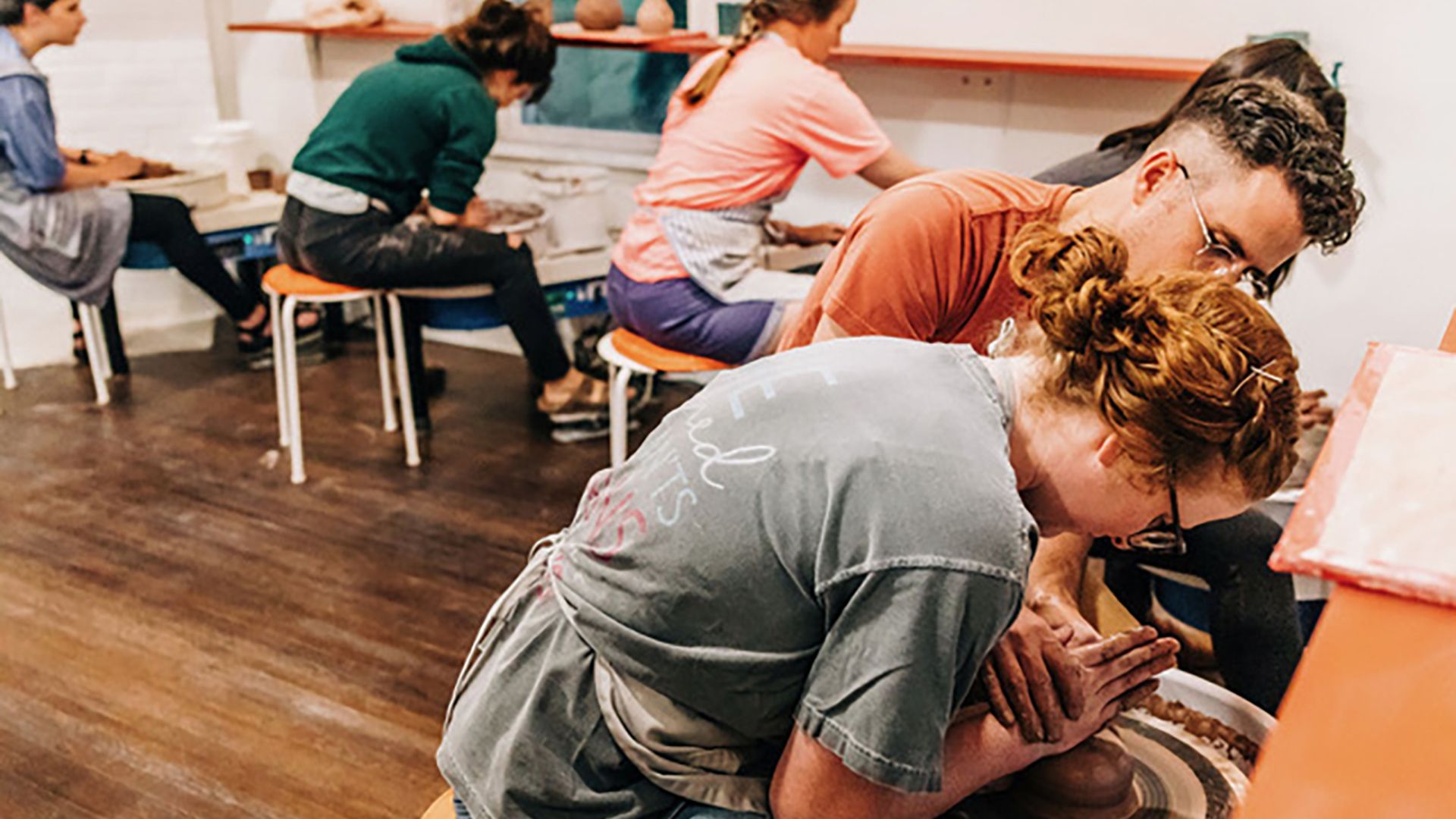 Several students sit on stools at pottery wheels in a bright studio. In the foreground, a person with red hair shapes clay on a spinning wheel as an instructor watches closely.