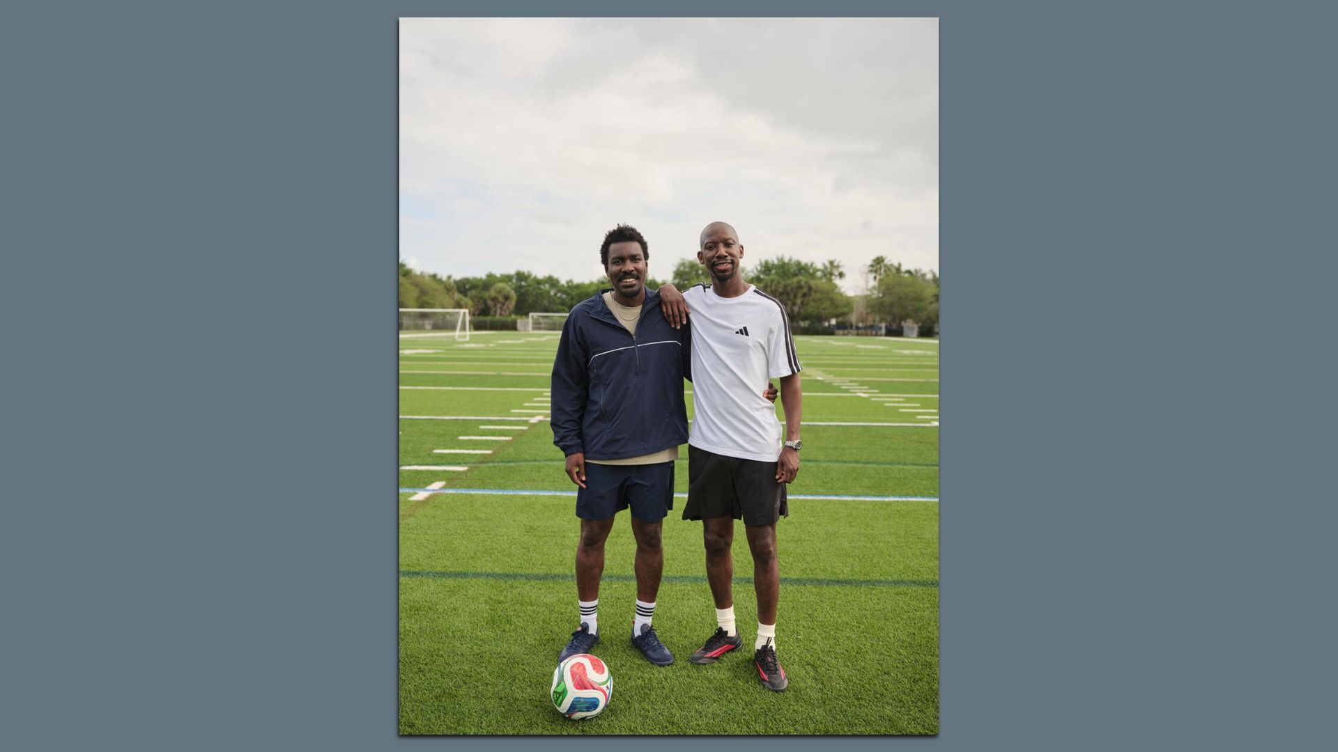 Two men pose with arms around on a green soccer field. 