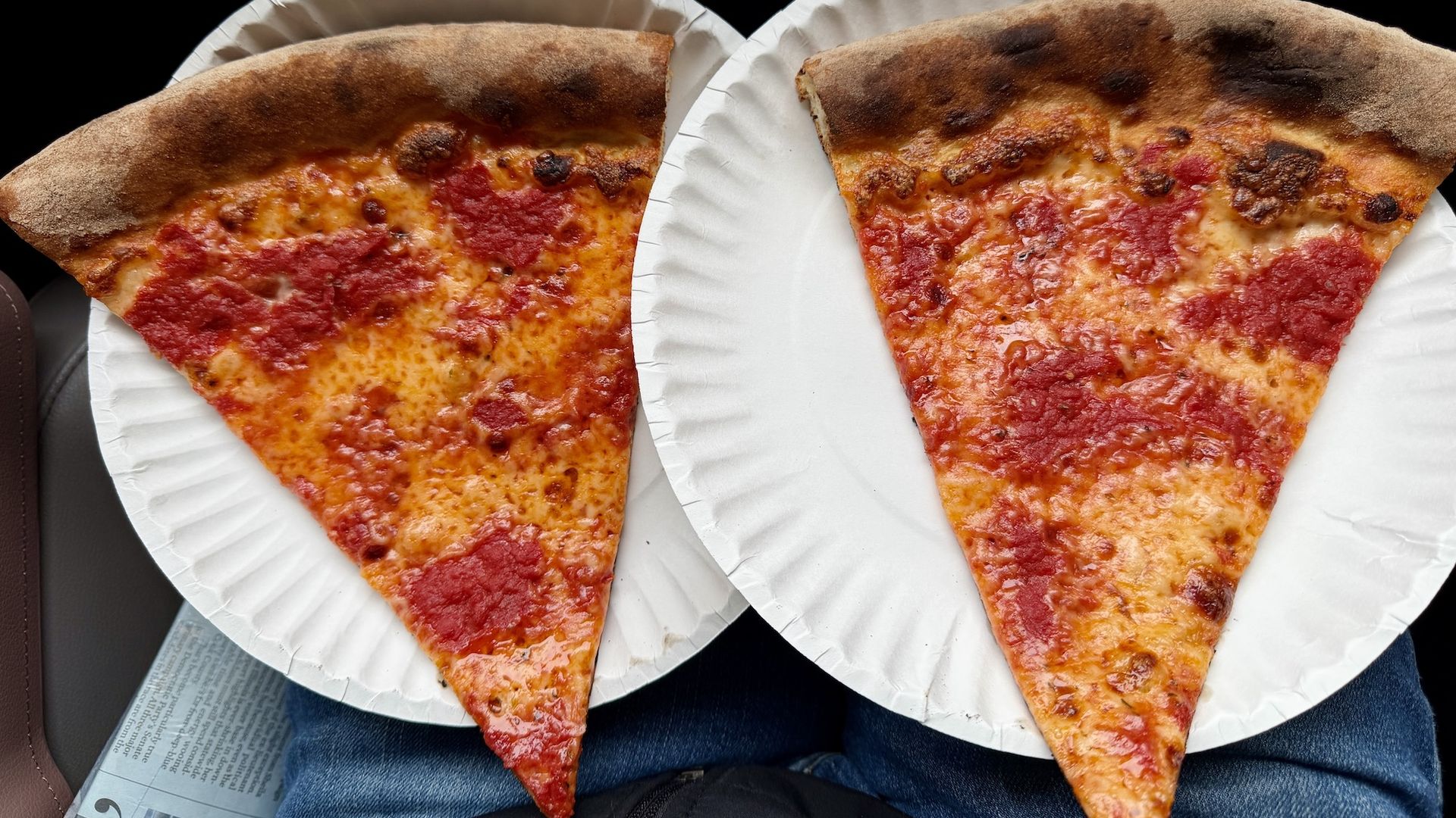 Two large slices of cheese pizza with tomato sauce on thin, slightly charred crusts, each on a white paper plate resting on a person's lap wearing blue jeans.