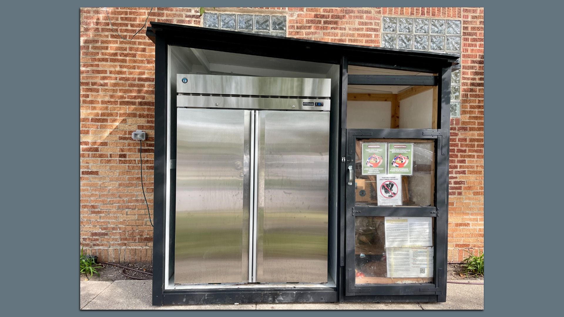 Stainless steel double-door refrigerator in a black-framed outdoor kiosk against a brick wall with glass blocks above. Notices and signs are posted on the right glass door.