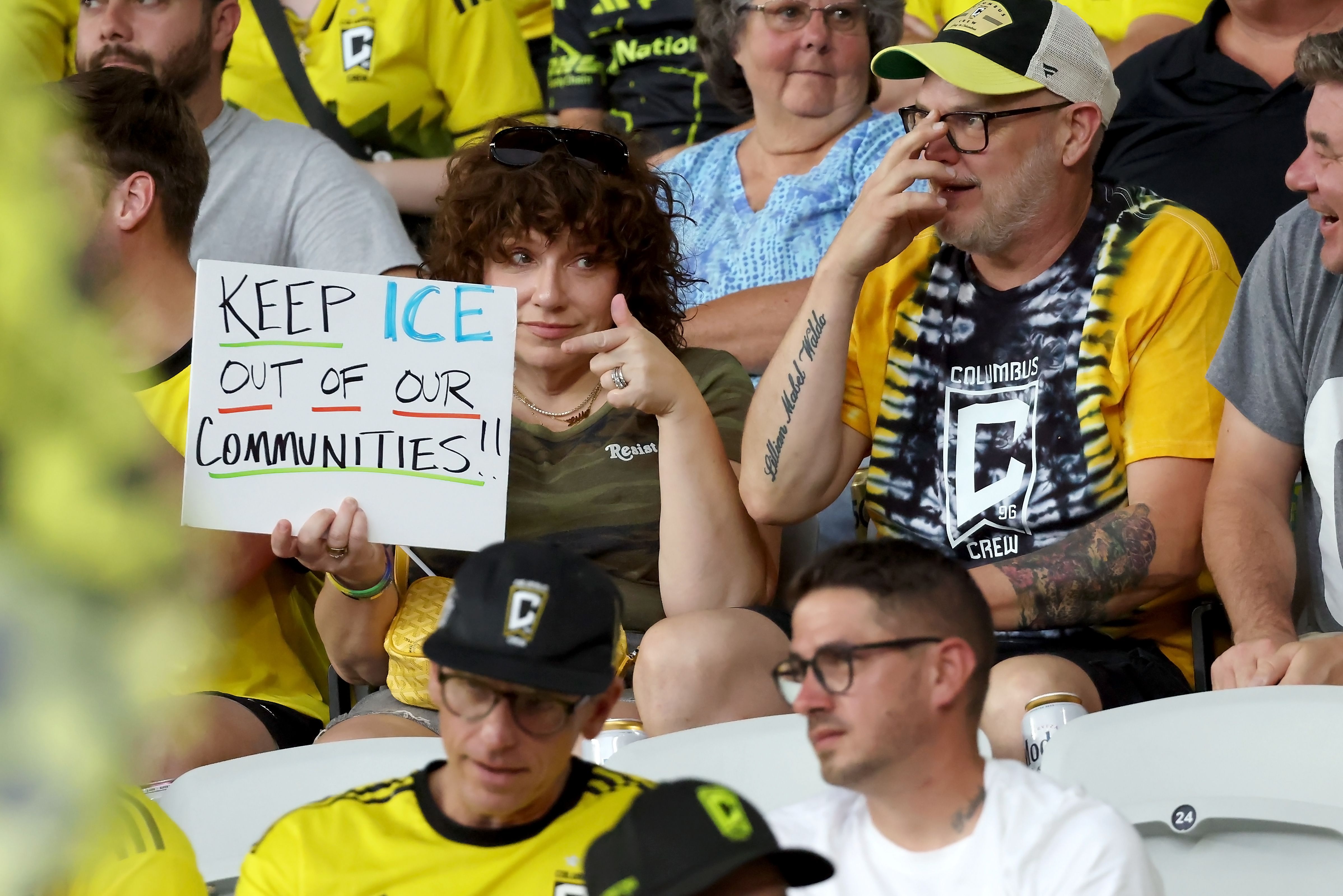 A woman with curly hair in a green shirt holds a sign saying "KEEP ICE OUT OF OUR COMMUNITIES!!" in the stands at a Crew match, surrounded by people wearing Columbus Crew apparel.