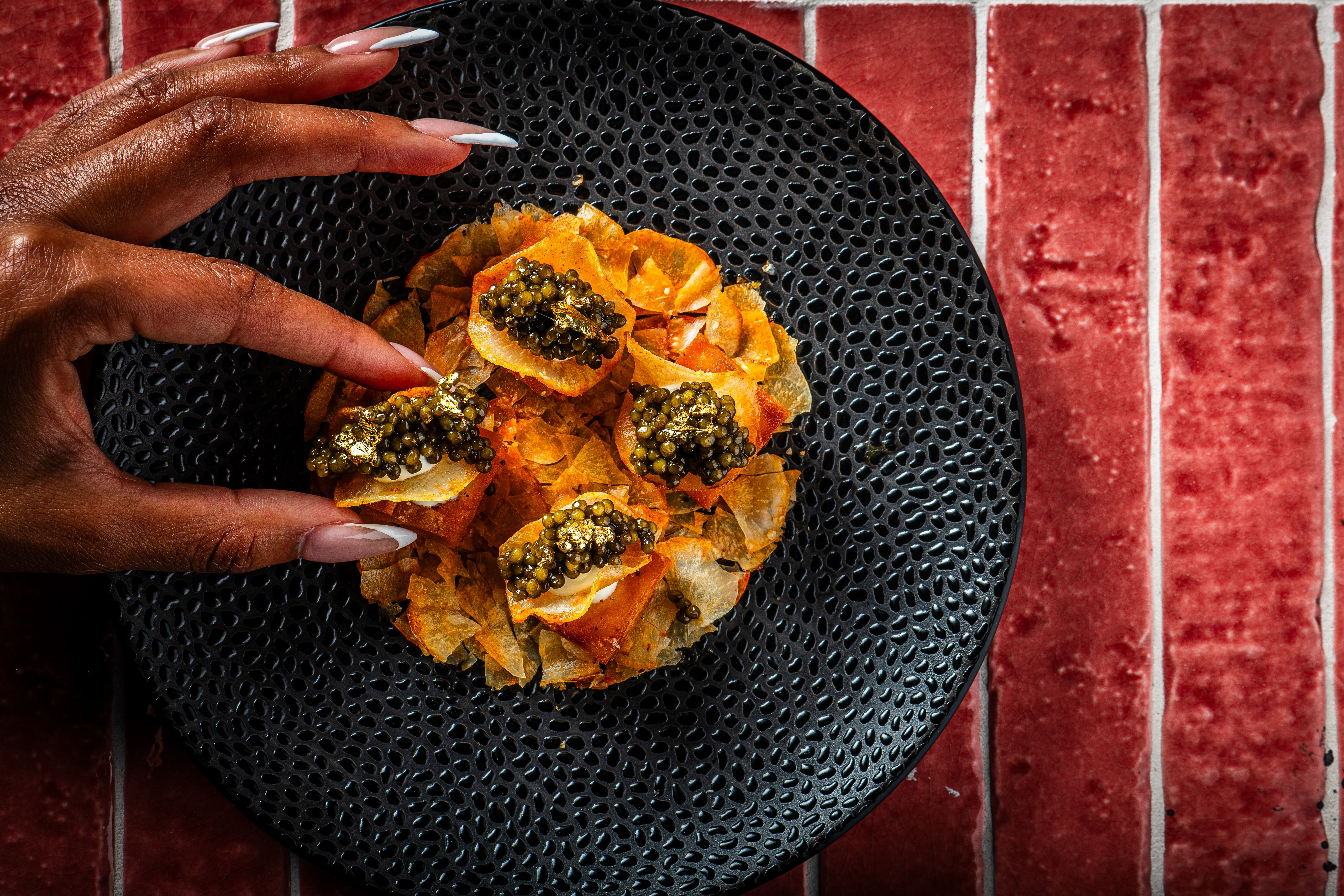 Close-up of a dark-skinned hand with long white nails resting on a black perforated plate of orange tortilla chips topped with caviar and gold leaf, set against a red brick background.