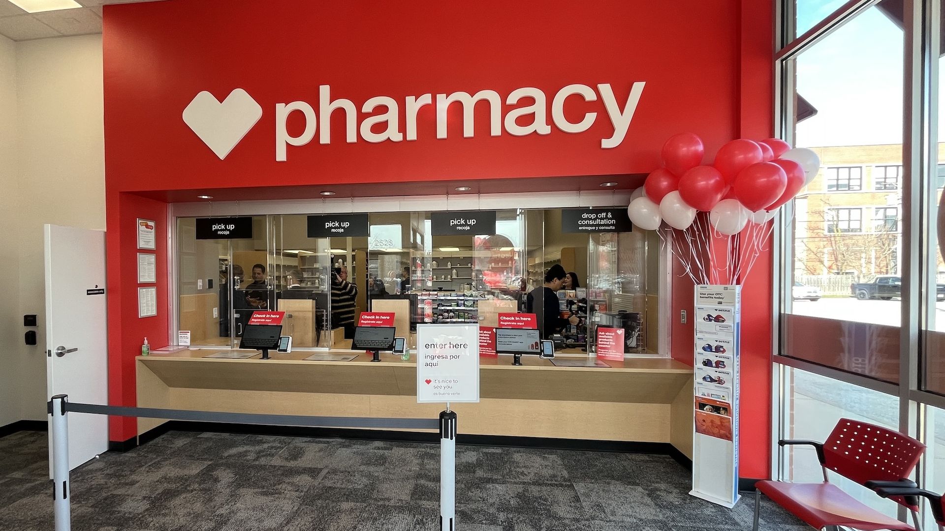 Interior of a bright pharmacy with a large red wall and a white heart icon beside the word pharmacy. A long counter with pickup windows, signs, and red-and-white balloons near glass walls.