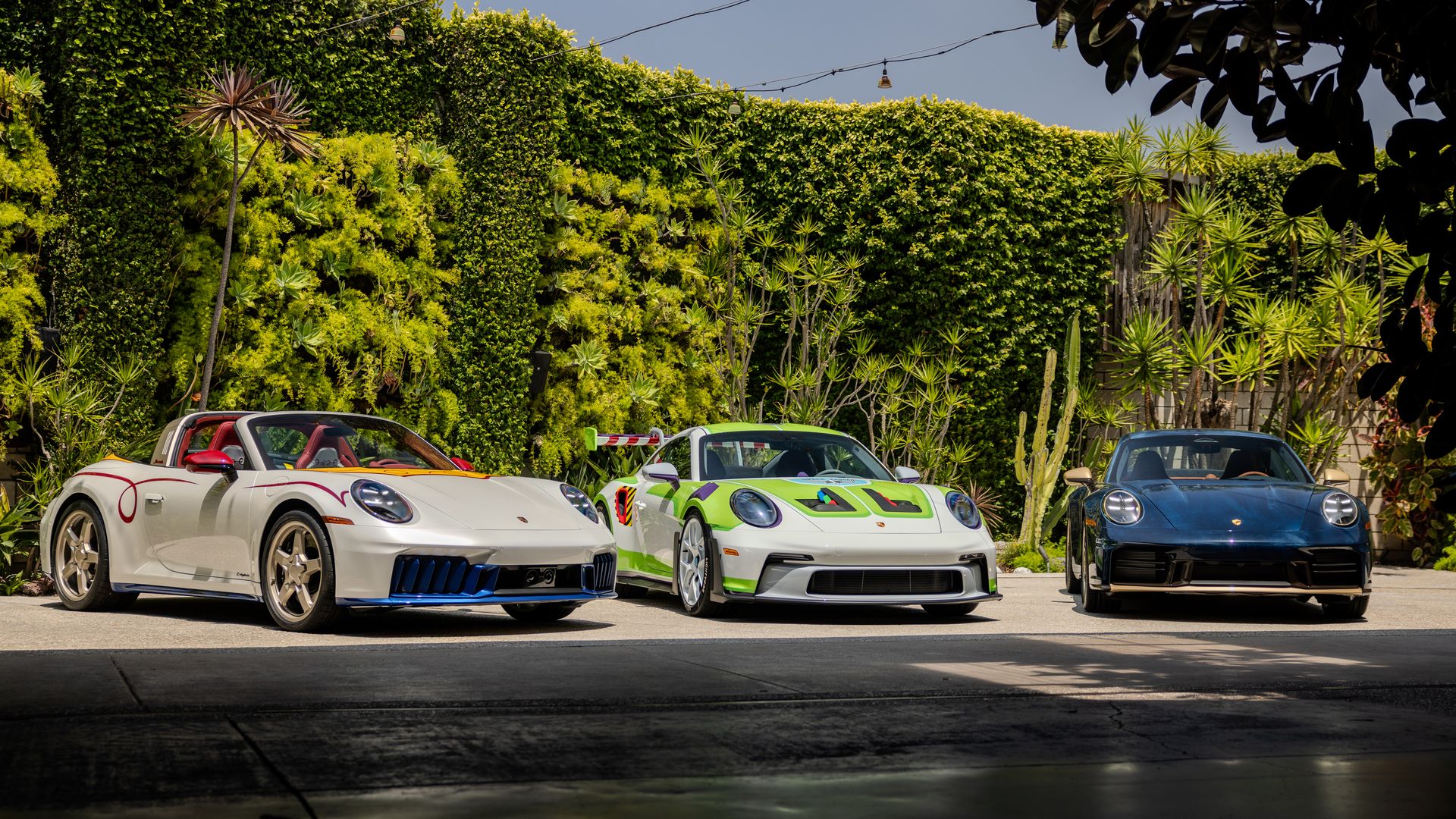 Three Porsche convertibles in white, green, and blue sit in a lot before a tall green hedge and tropical plants; string lights stretch overhead under a clear blue sky.