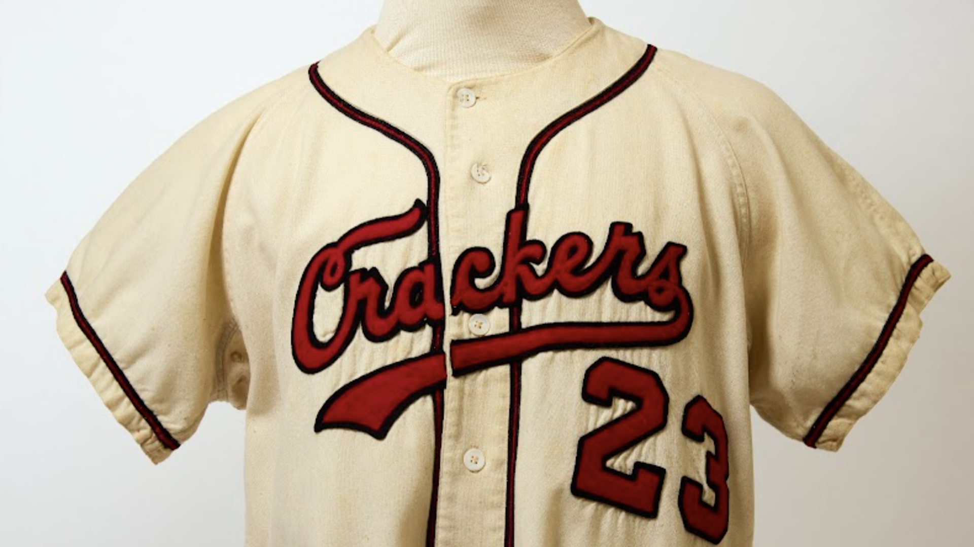 A cream-colored vintage baseball jersey on a display mannequin, featuring red-outlined script "Crackers" across the chest and the red number 23, with red piping along the collar and sleeve edges.
