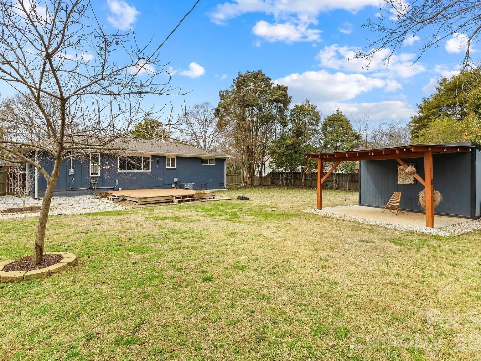 Backyard with a blue house on left, wooden deck, leafless tree in foreground, grass lawn, and a wooden pergola with single chair under a partly cloudy blue sky.