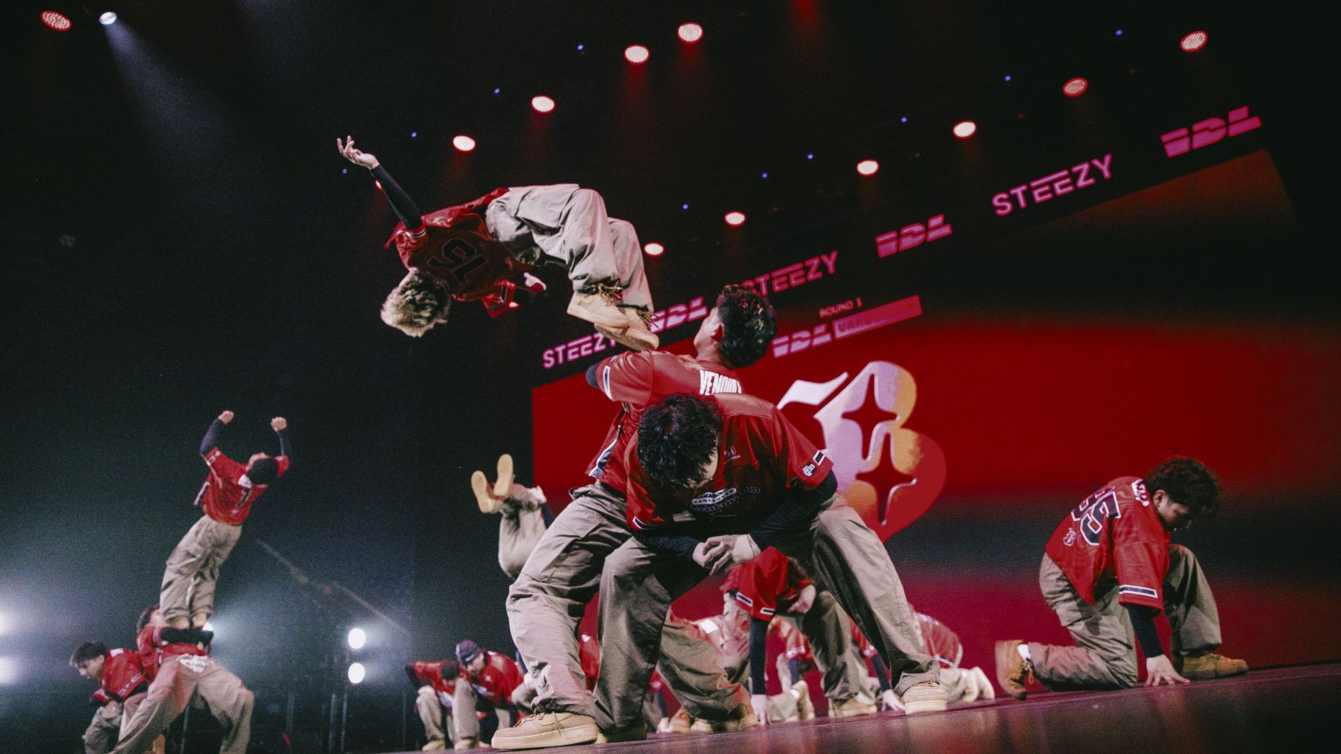 Group of dancers in red jerseys leap mid-air on a stage under red and blue spotlights, with a red backdrop displaying STEEZY logos for a high-energy performance.