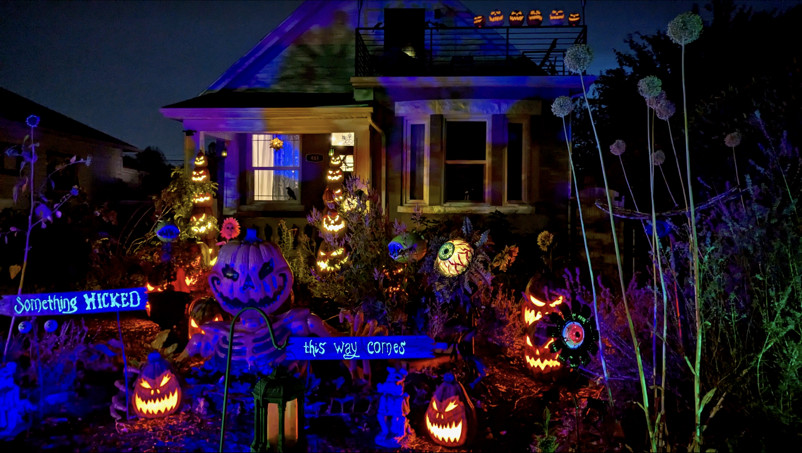 Nighttime Halloween yard with glowing jack-o'-lanterns along the porch, blue and purple lights, tall flowering stems, and a sign reading "Something Wicked This Way Comes".
