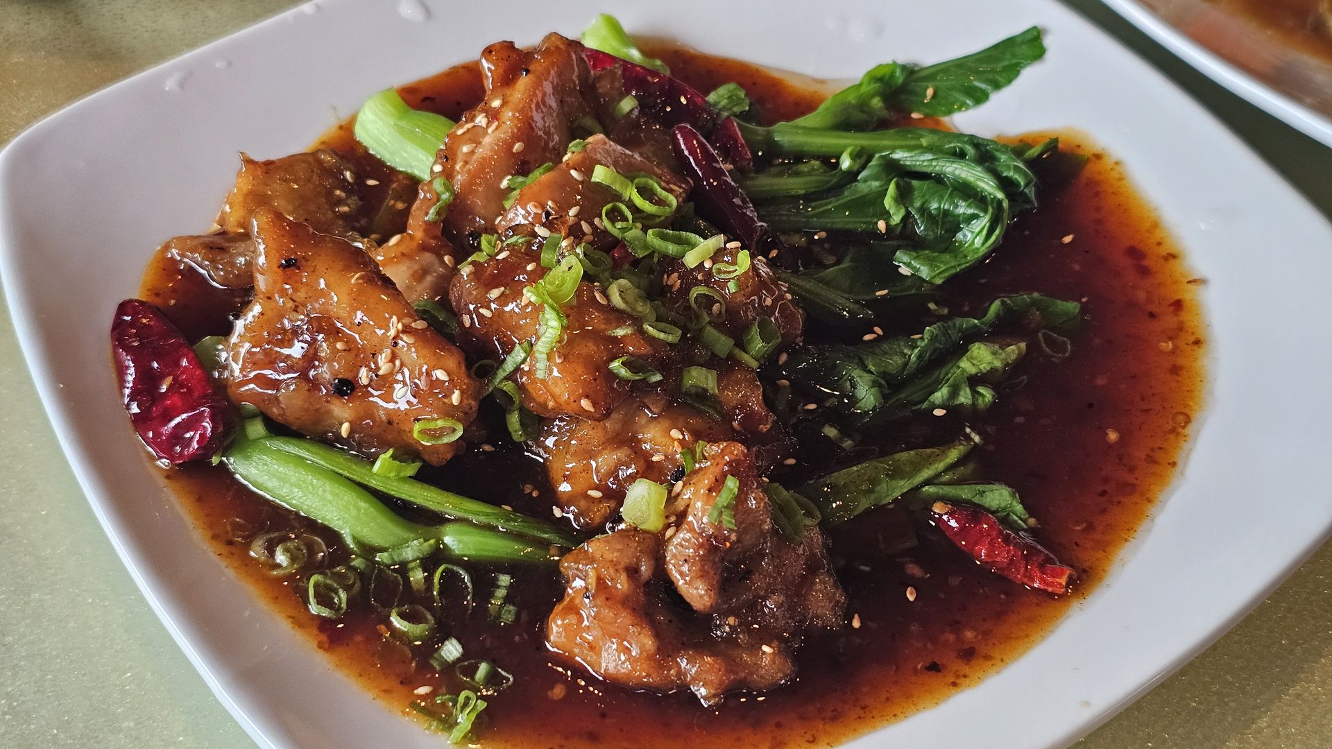 A white bowl of braised chicken in a spicy red sauce, topped with sesame seeds and sliced green onions, with wilted greens and dried chilies in the sauce.