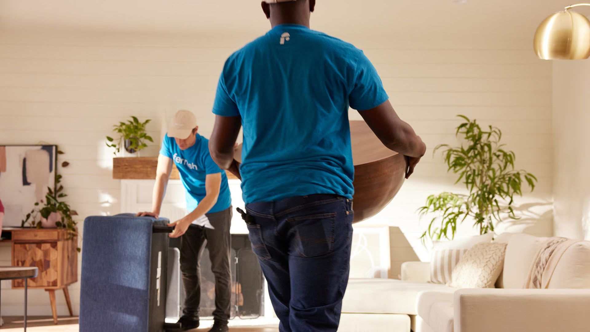 Two male movers, both in blue shirts, assemble furniture in a living room area with sand-colored carpet.