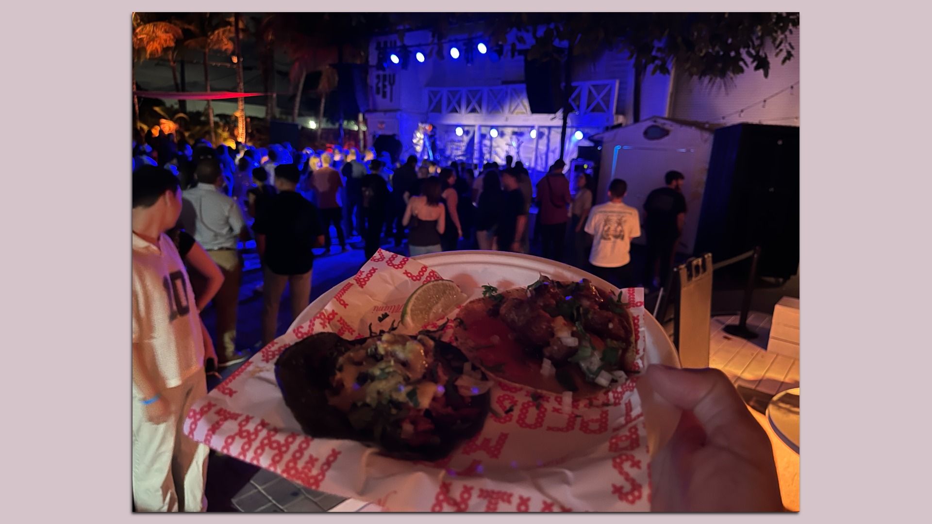 Hand holding a plate with two tacos and a lime wedge in front of a crowd at a nighttime outdoor concert with blue stage lights.