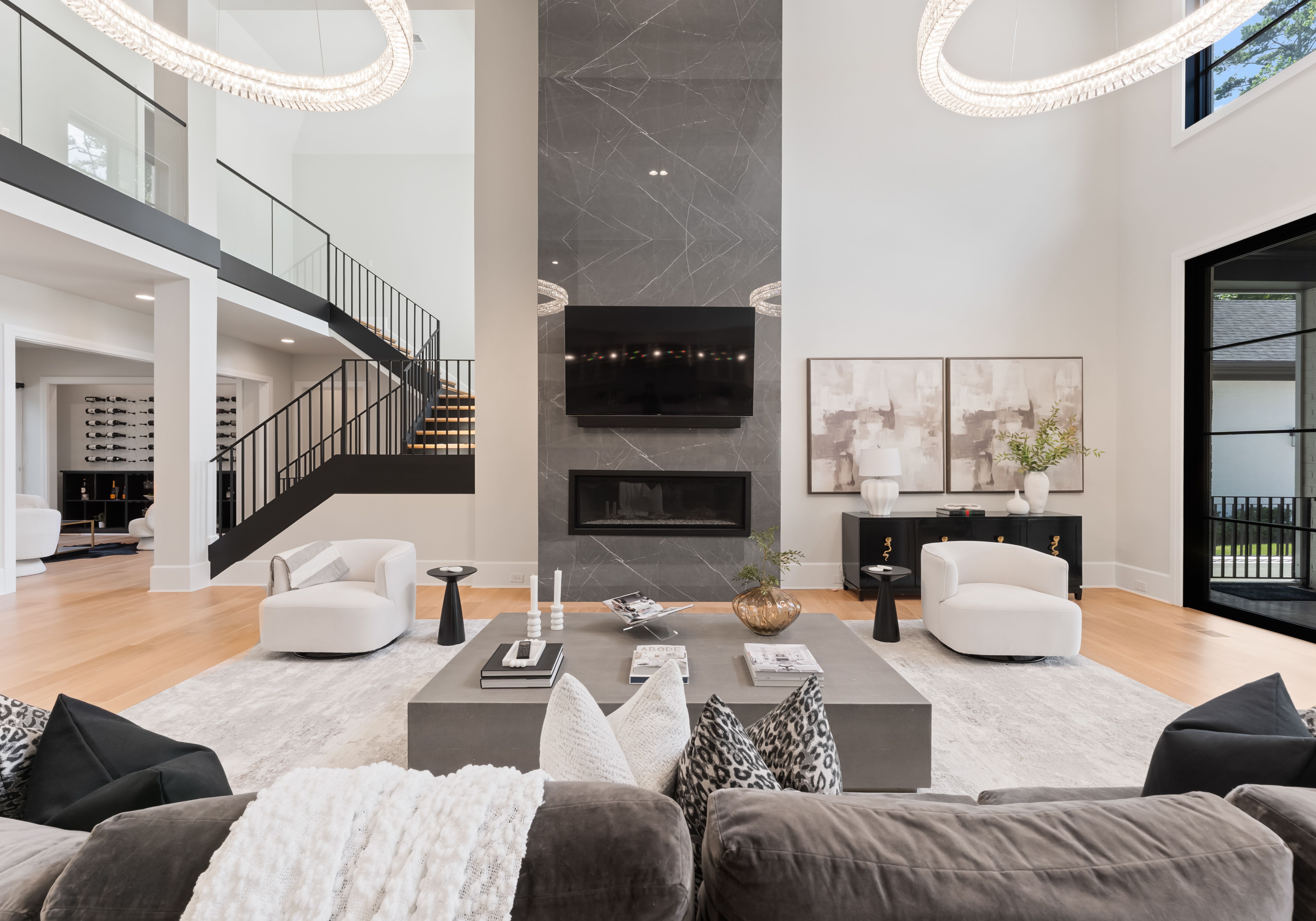 Modern living room with high ceilings featuring a large gray marble fireplace, wall-mounted TV, two white curved chairs, black side tables, neutral artwork, and light wood floors.