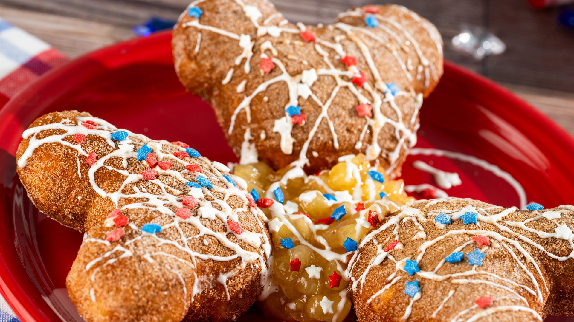 Close-up of golden-brown fried doughnut holes on a red plate, drizzled with white icing and red, white, and blue star sprinkles; a wooden table forms the background.