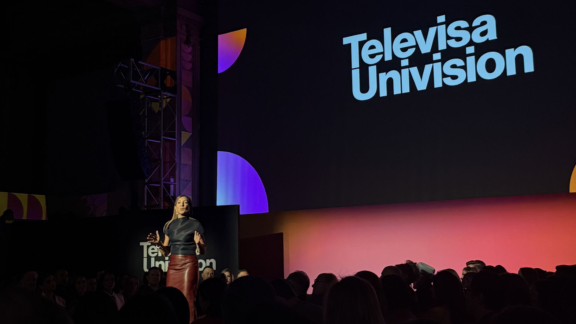 Donna Speciale speaking in front of a seated crowd with the name TelevisaUnivision displayed behind her