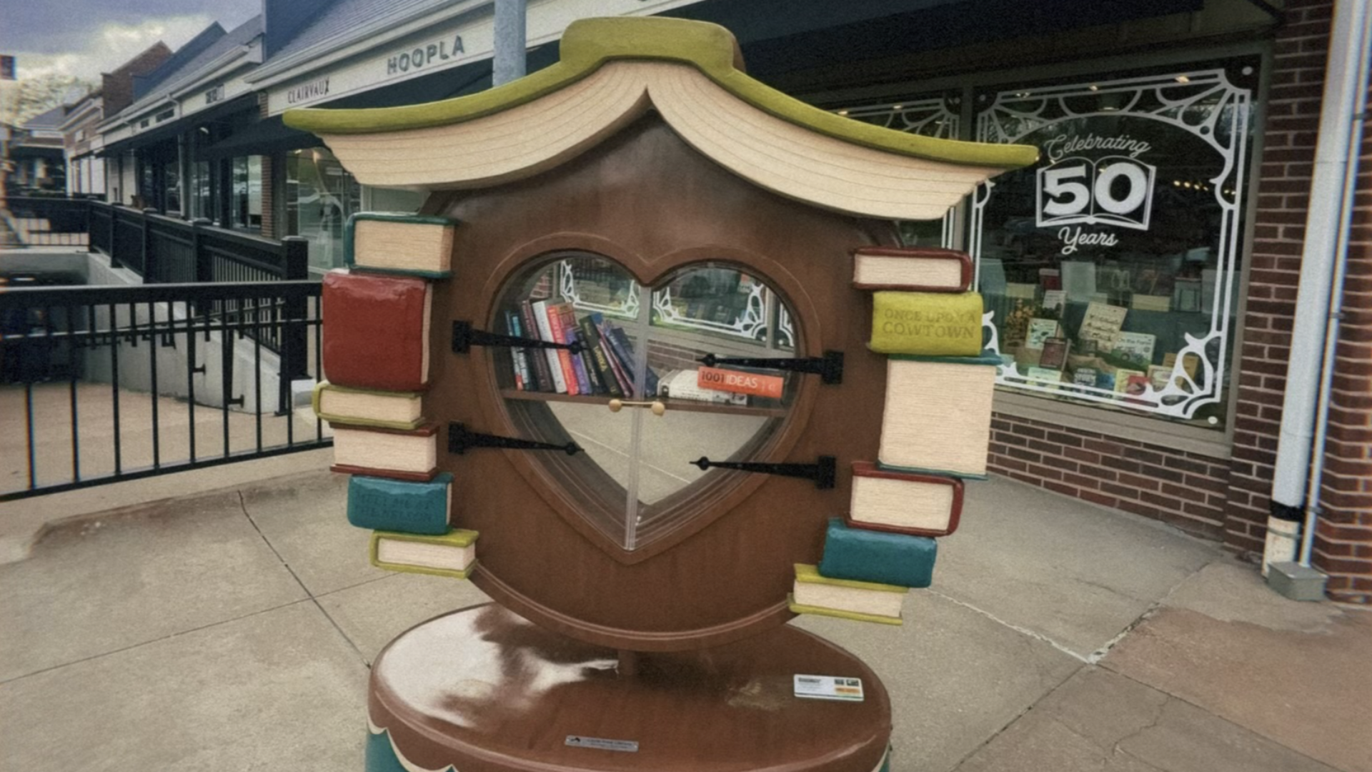 Whimsical giant book sculpture on a sidewalk: a circular glass window filled with books, stacks of colorful books on both sides, and a curved roof; storefronts and a Celebrating 50 Years window in the background.