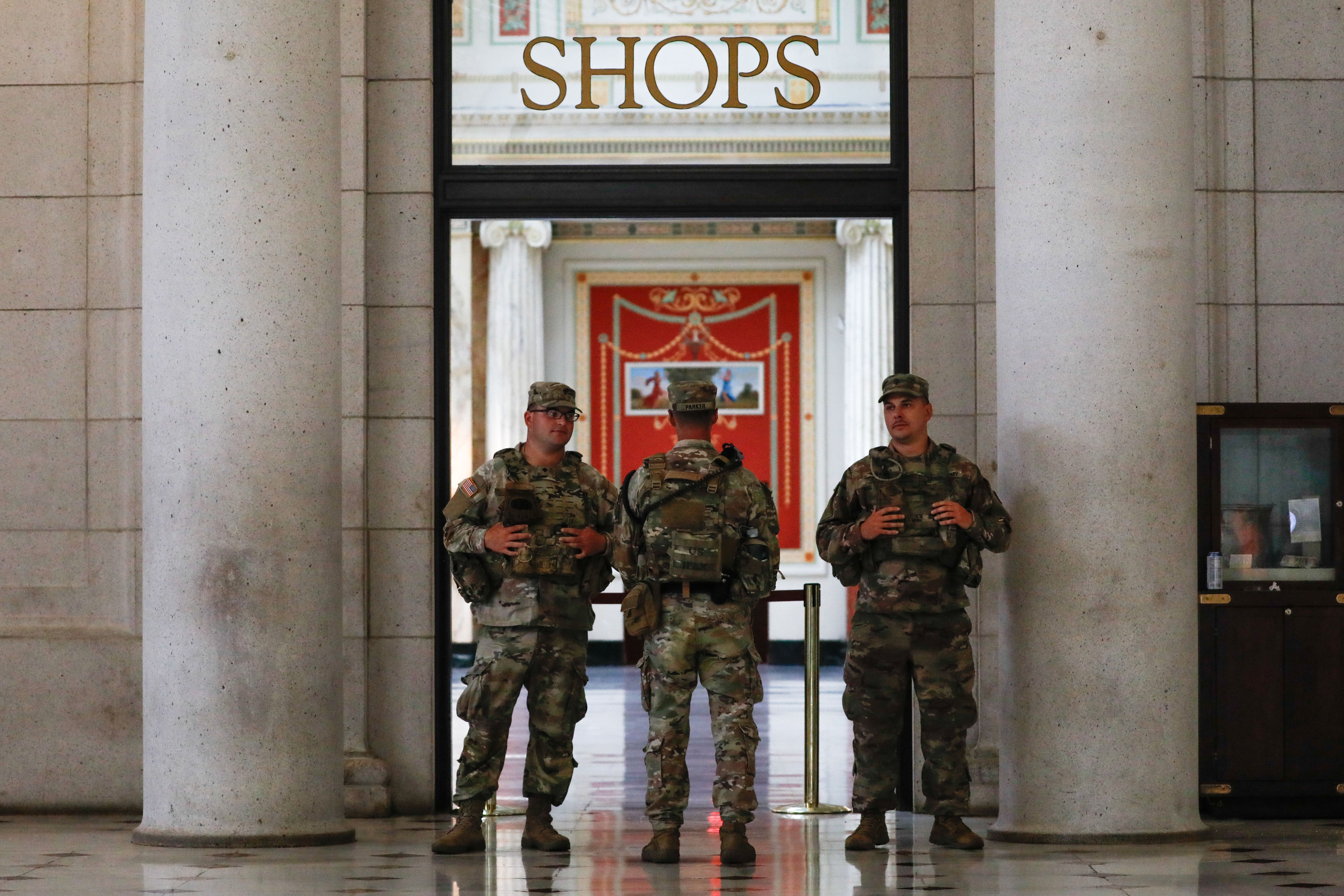 Three soldiers in camouflage uniforms standing guard in a marble building entrance beneath a sign reading "SHOPS." Decorative red and white interior visible in the background.