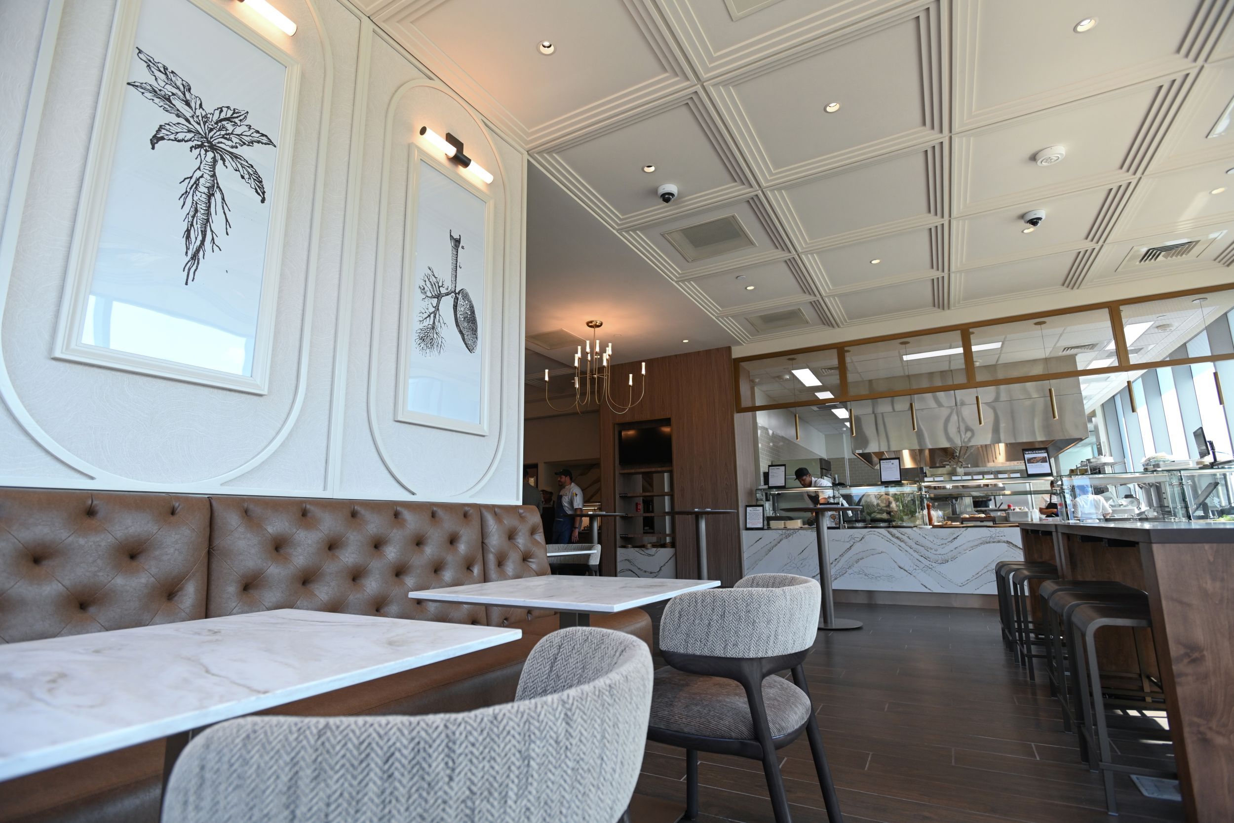 Inside a modern cafe: brown tufted leather banquette along white walls with framed black botanical prints; marble tables; chandelier; geometric ceiling panels; open kitchen with glass counter.