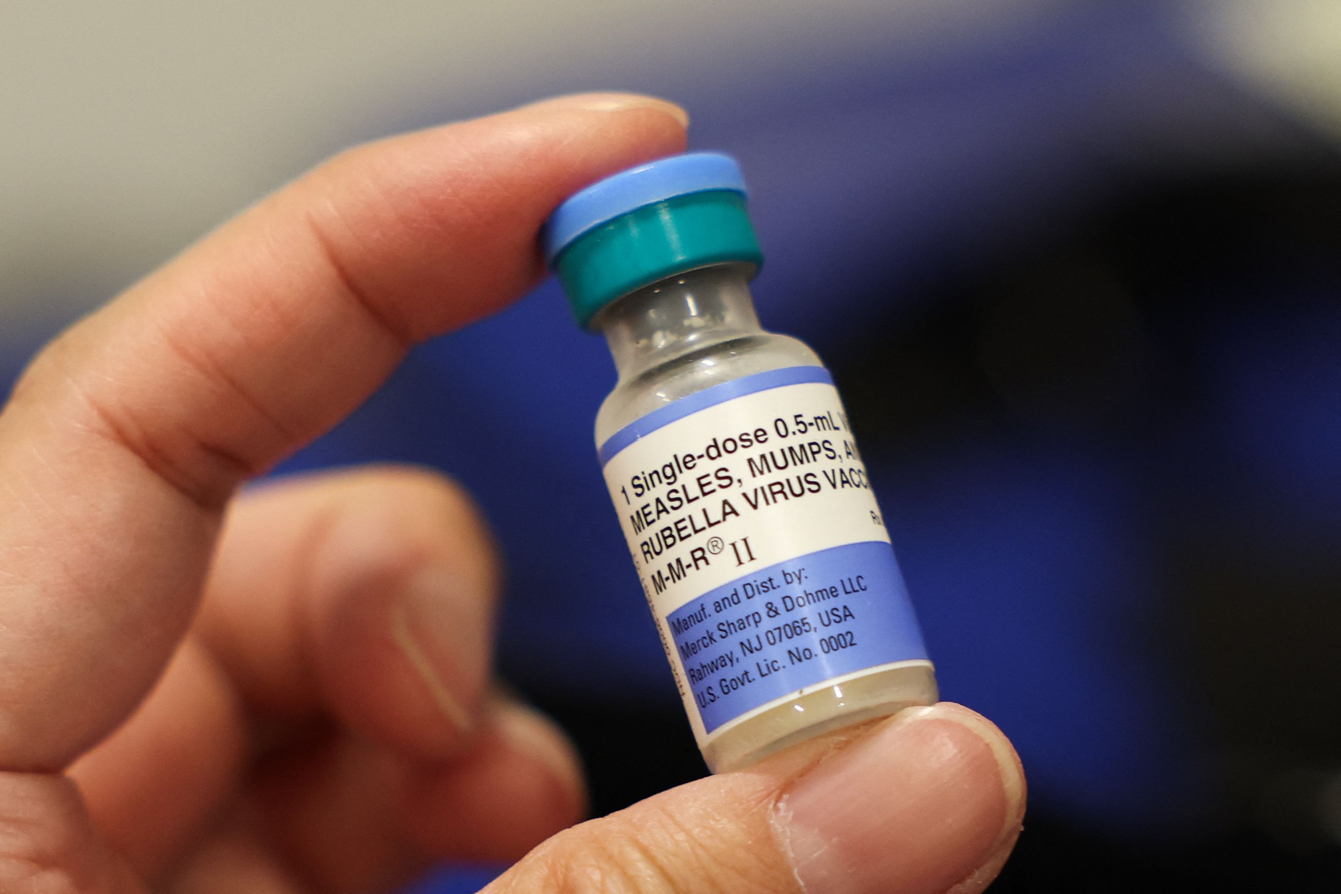 A small glass vial labeled “MMR” for measles-mumps-rubella vaccine held in gloved hands.