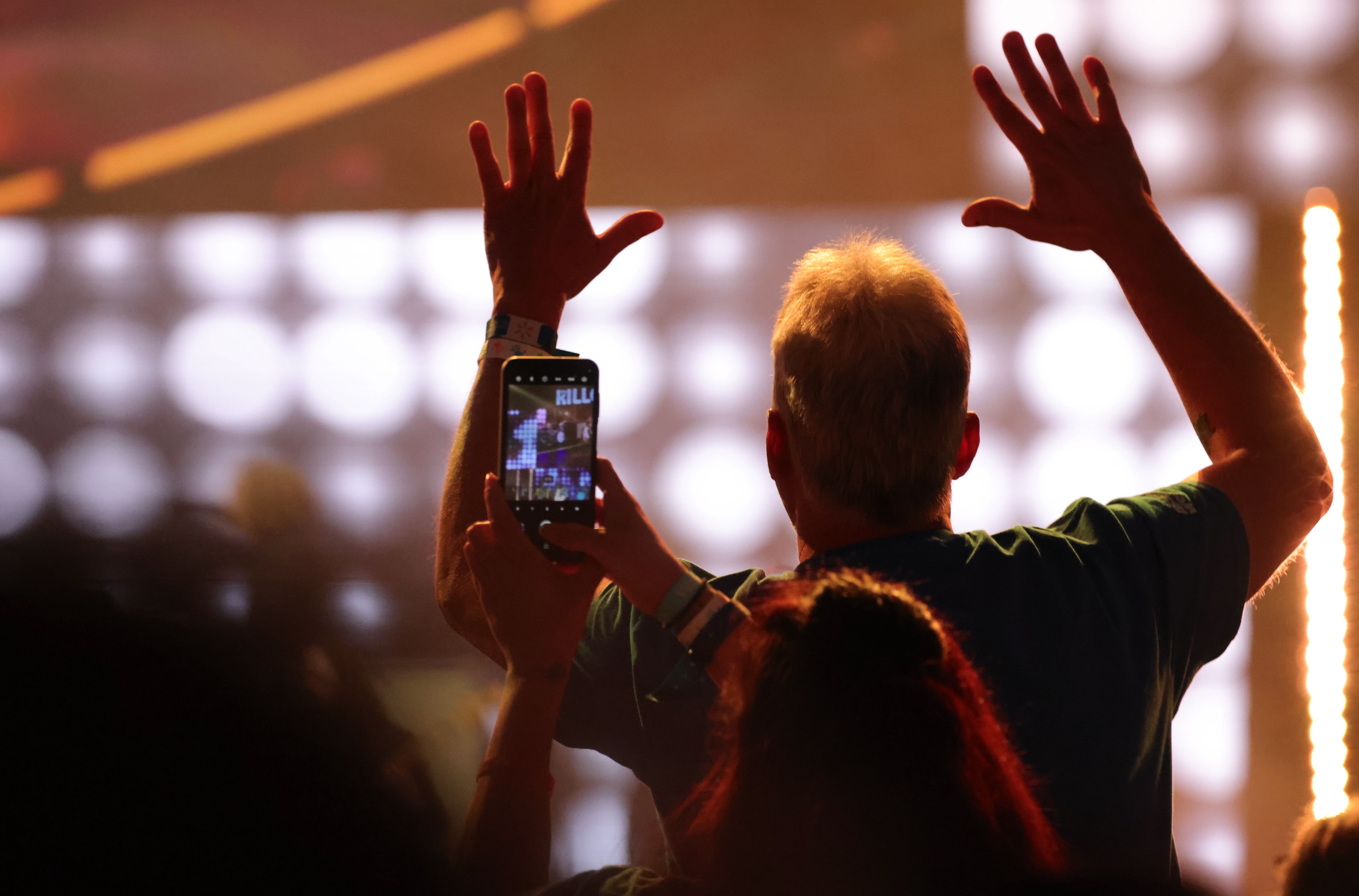 Man with hands raised at a concert while someone films the stage on a phone amid bright, blurred lights.