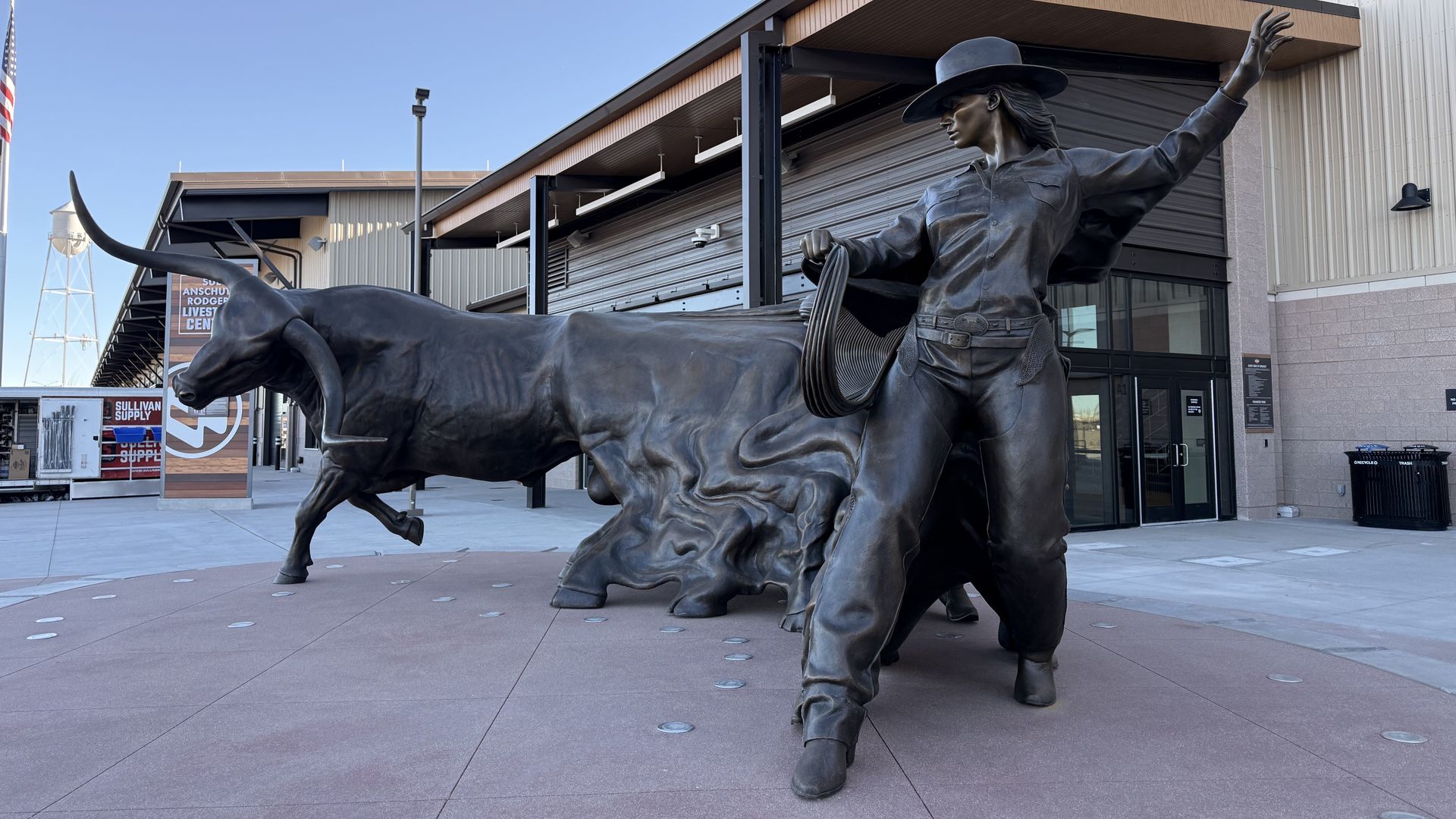 Bronze statue of a cowgirl in a hat holding a lasso beside a longhorn steer outside a building under clear blue sky.