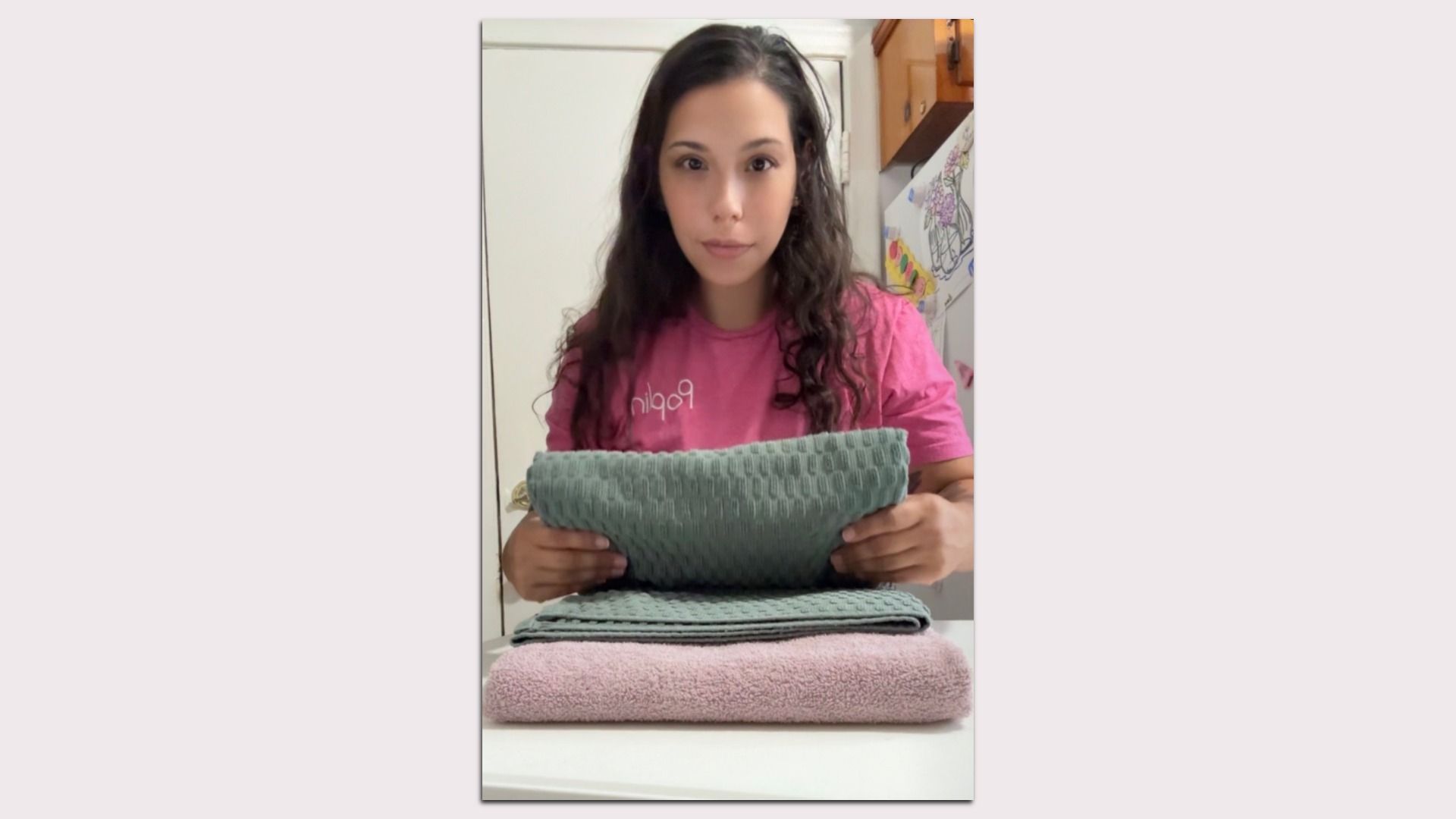 Young woman with long dark hair in a pink shirt holds a teal towel, while stacked towels lie on a table; kitchen background shows a decorated fridge.