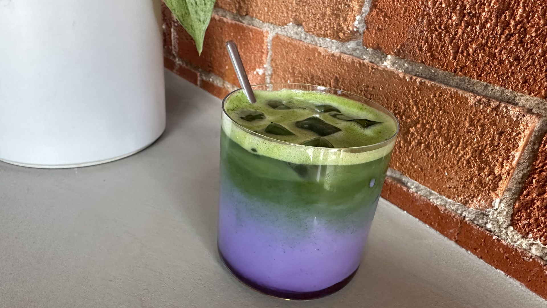 A tall glass with a purple-to-blue-to-green gradient contains a green iced drink with dark ice cubes; a metal straw sticks out. The glass sits on a grey surface beside an orange brick wall.