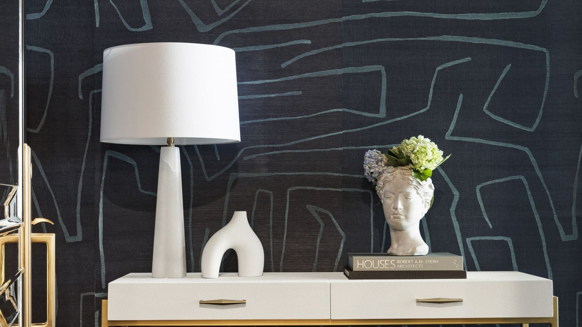 A photo showing a black wallpaper with chalky white modern lines through it. In front of it is a side table with a lamp, vase shaped like a head and a modern sculpture. A rectangular stool with a comfy seat is also visible.