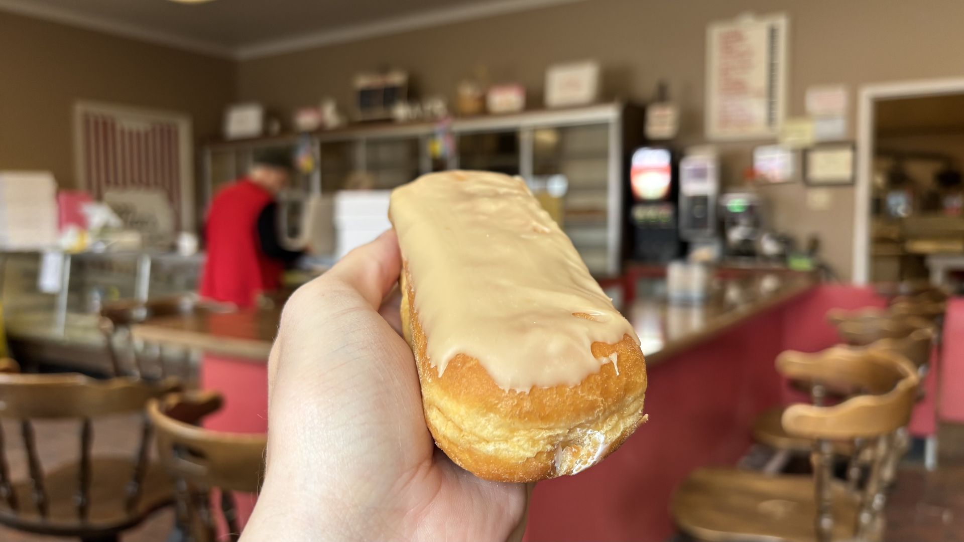 A hand holds a long, glazed doughnut in a retro donut shop with a pink counter, blurred background, and a person in red working behind the counter.