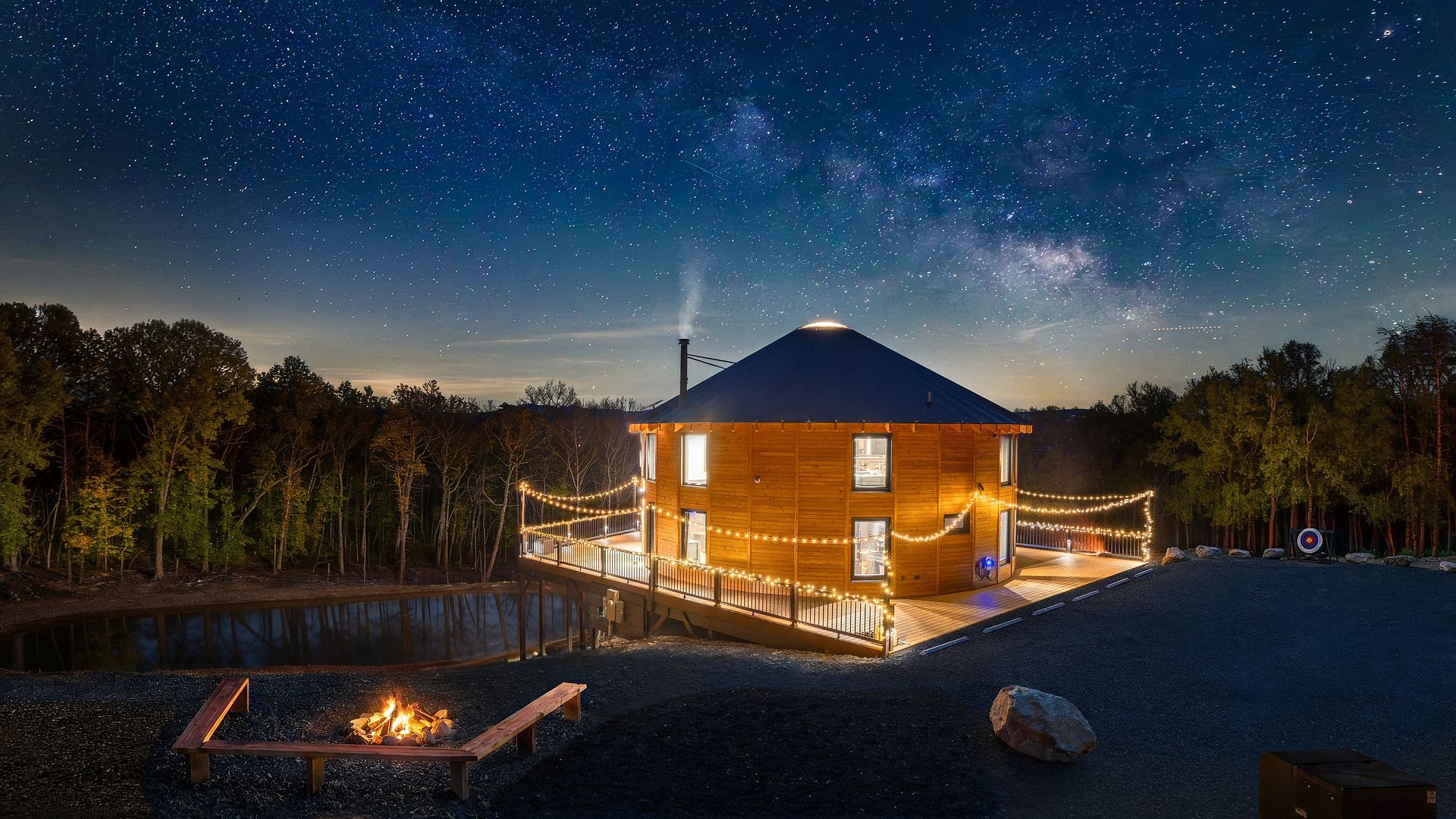 Night scene of a wooden round house with a cone roof, warm string lights around the deck, a fire pit in the foreground, and a star-filled sky with the Milky Way overhead; trees line the background.