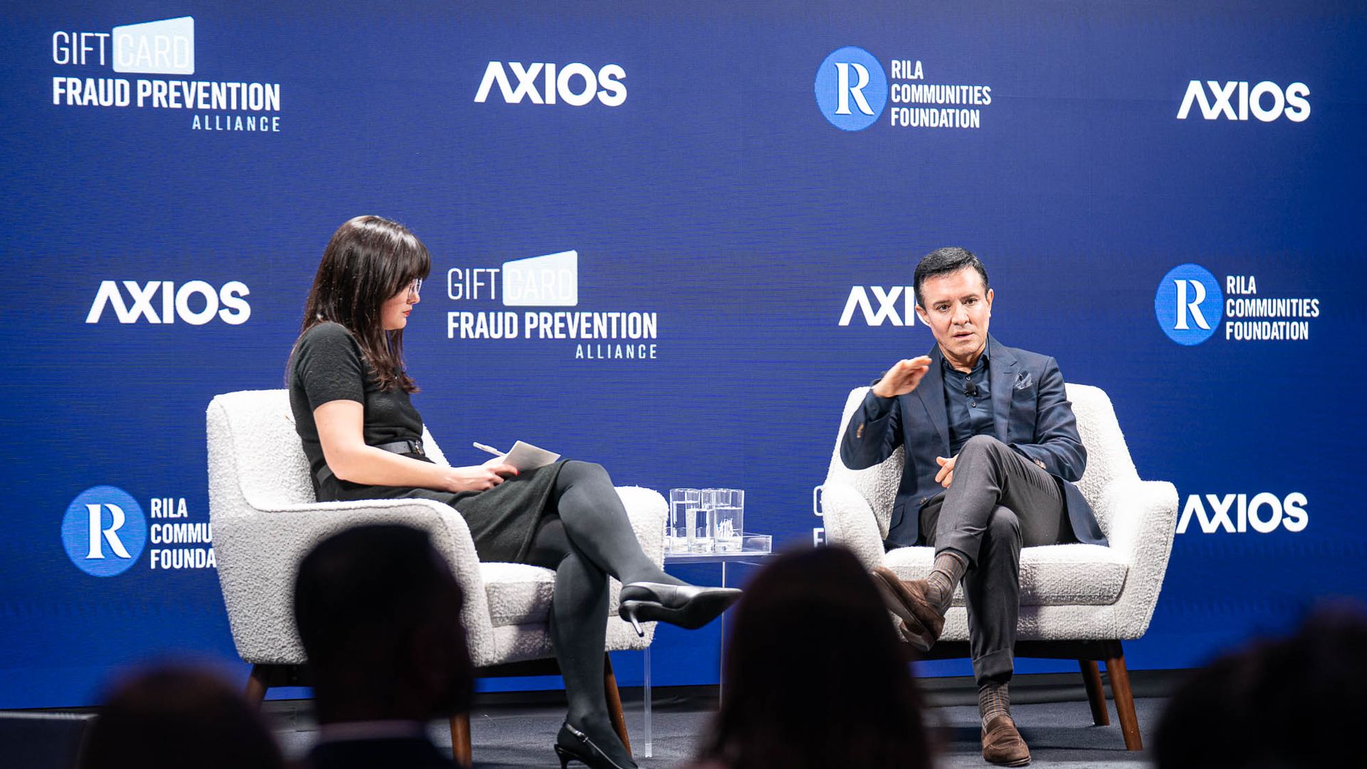 Two people seated in white armchairs on a stage with a blue backdrop featuring logos for Gift Card Fraud Prevention Alliance, Axios, and RILA Communities Foundation, engaged in discussion.