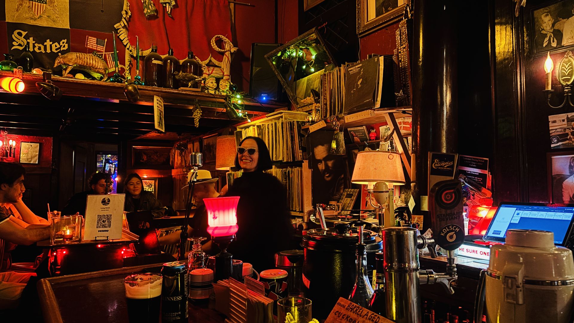 Cozy, dimly lit retro bar full of knickknacks and records. A smiling woman wearing sunglasses stands by a vintage microphone; patrons sit at the bar with laptops and drinks amid warm amber glow.
