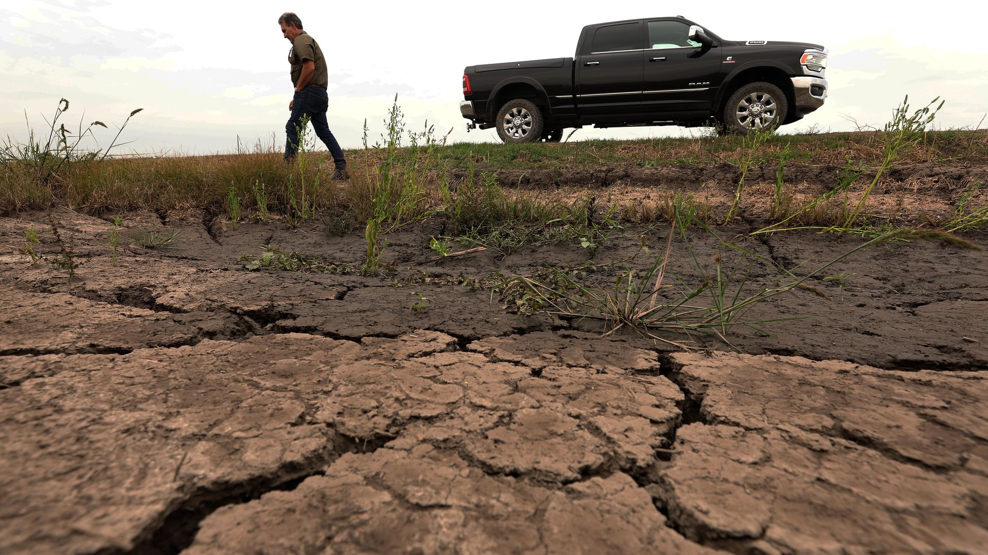 Photo shows a man walking near dry, cracked dirt with a truck in the background.