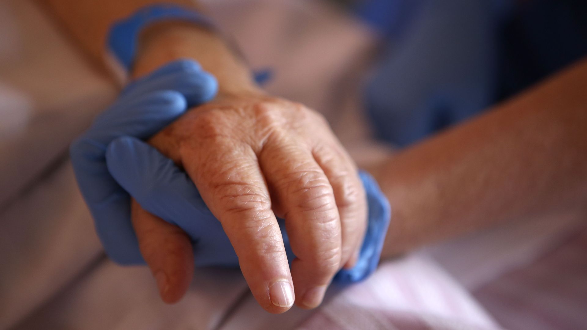 A nurse wearing protective gloves holds the hand of a patient in palliative care.