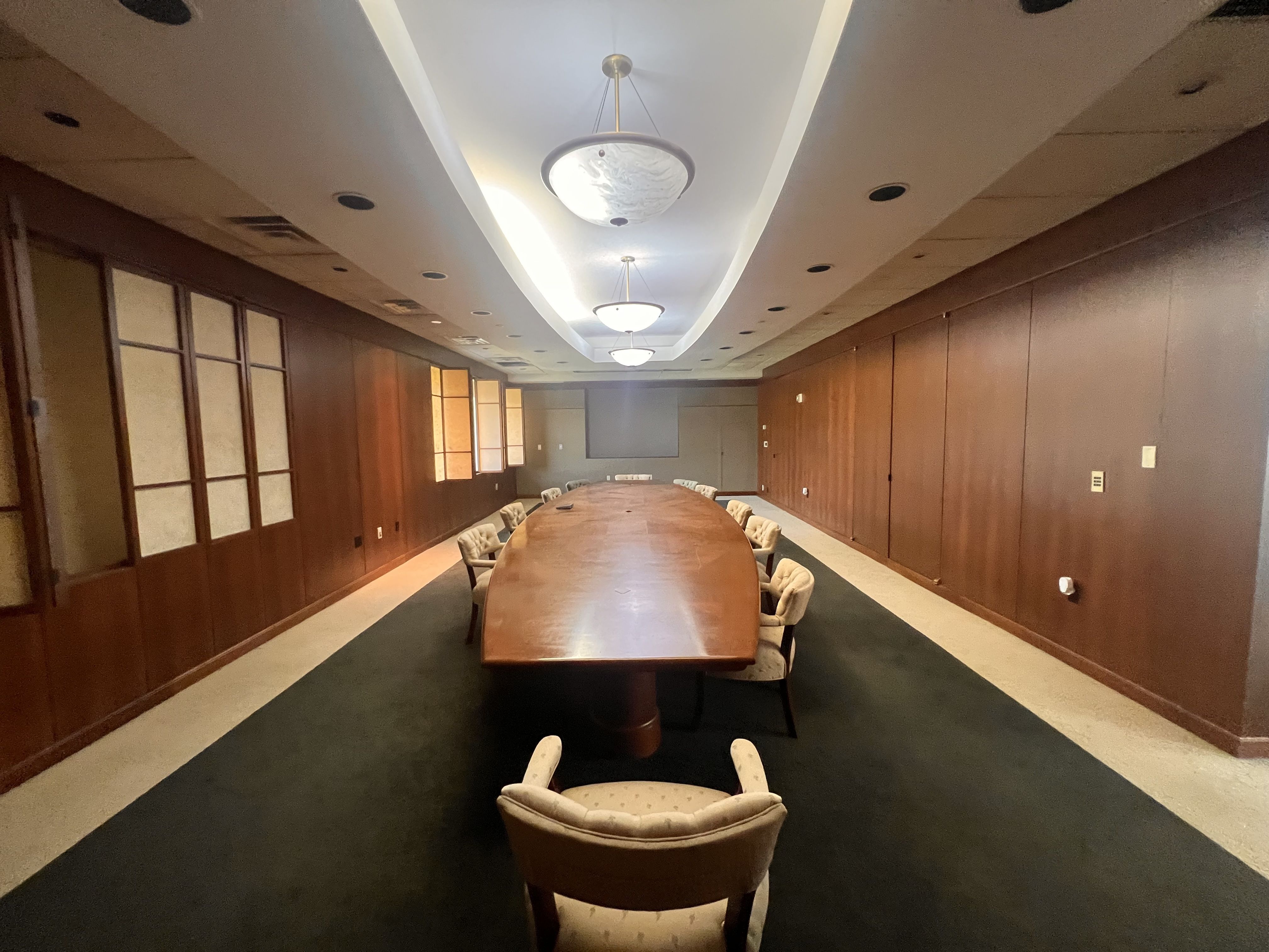 Long wood conference table with upholstered chairs in a wood-paneled meeting room. White curved ceiling lights hang above, flanked by dark carpet and wall panels, creating a formal office setting.