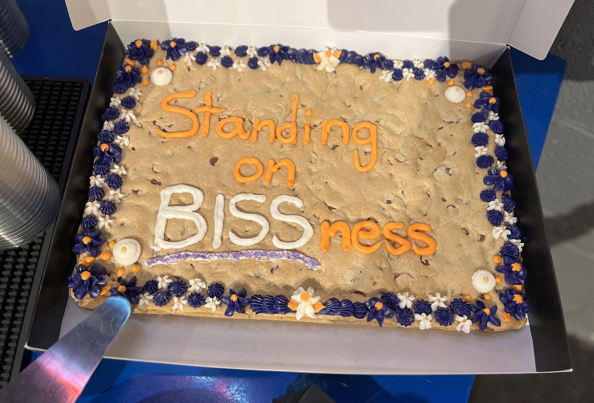Rectangular cookie cake in a white box with purple and white border. Beige icing with chips. Orange icing forms the words Standing on and mess; white icing creates BISS in the center with a purple underline.