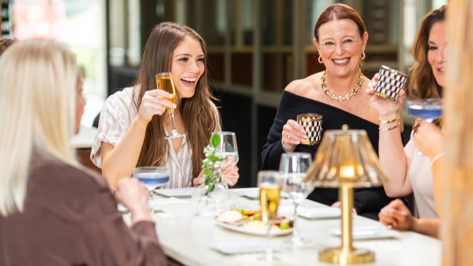 A group of women sit around a marble table in a restaurant, smiling and toasting with gold-rimmed glasses. Food, a small bouquet, and warm lighting are visible.