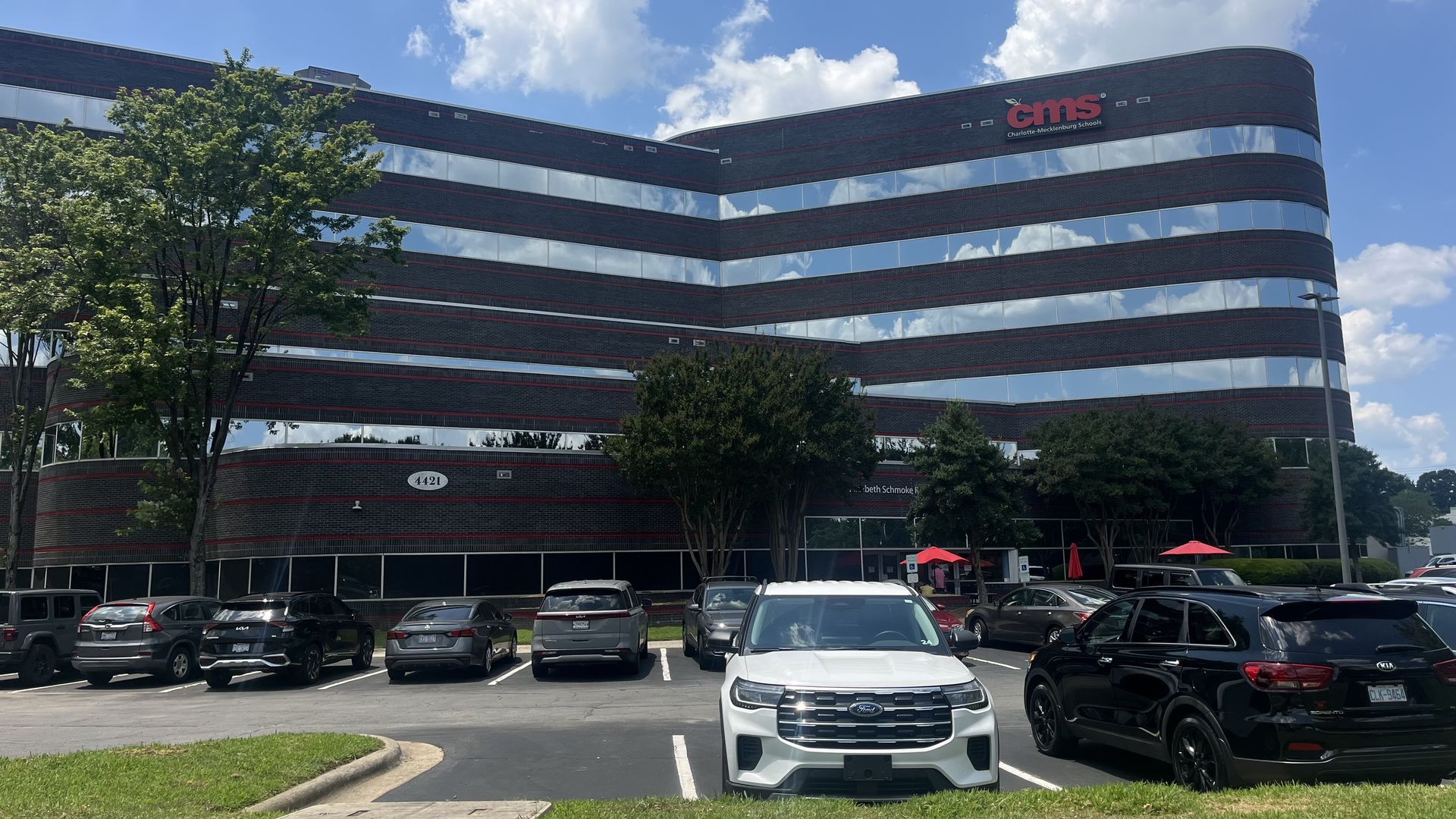 Modern curved glass-and-brick office building with red horizontal bands, reflecting a blue sky. A parking lot in front shows a white SUV in the center and several other cars beside green trees.