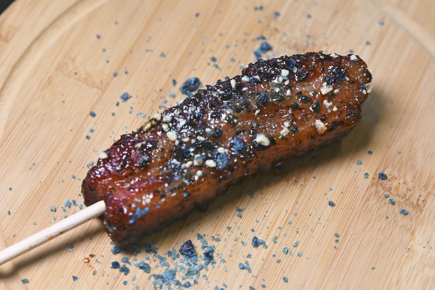 Close-up of a glazed grilled sausage on a wooden skewer, coated with dark brown glaze and coarse salt/seed crystals, with blue specks scattered on a light wooden cutting board.