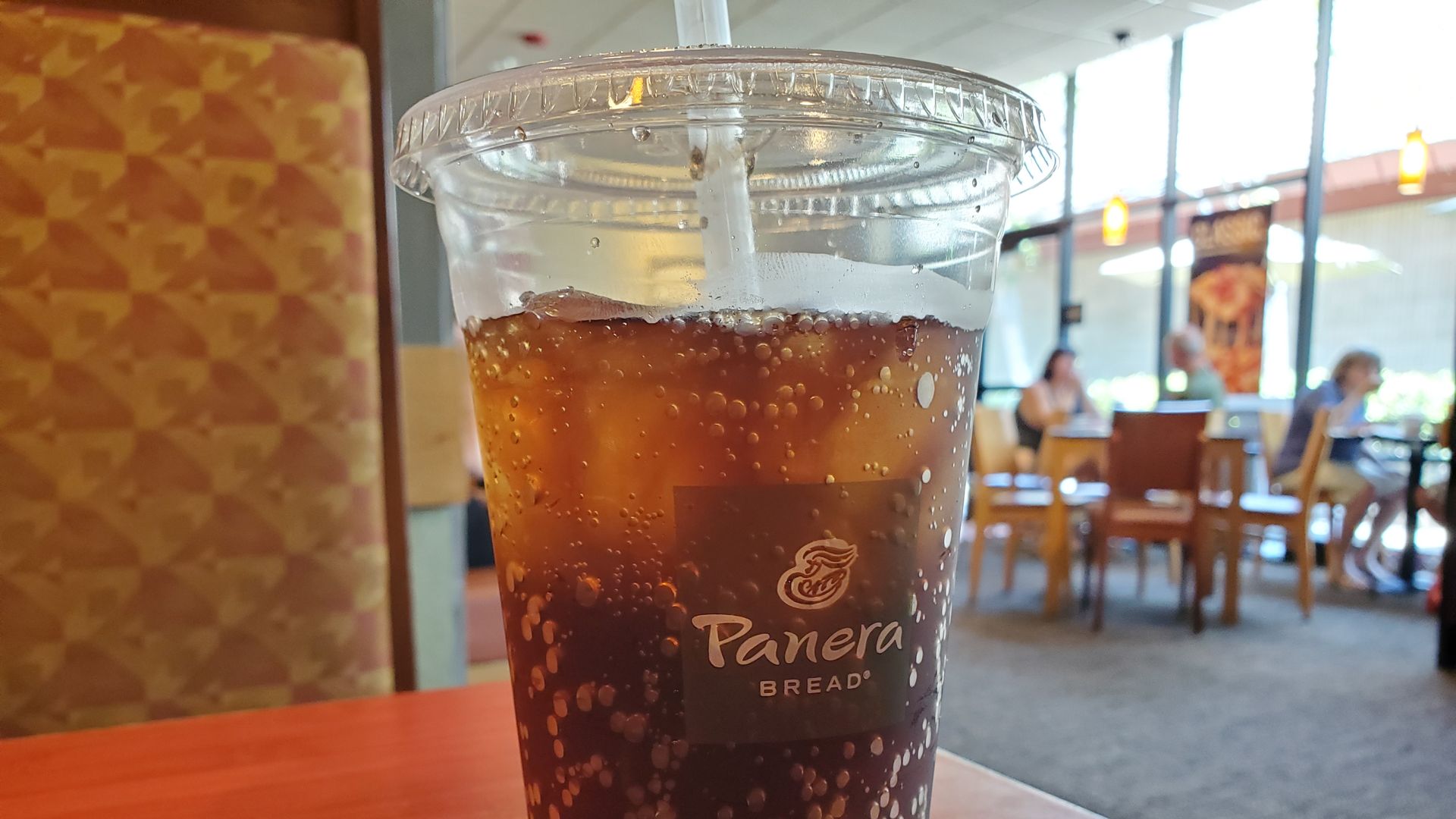 a clear plastic cup sits on a red countertop. It contains a dark soda and is covered in condensation. A straw is inserted into the lid.