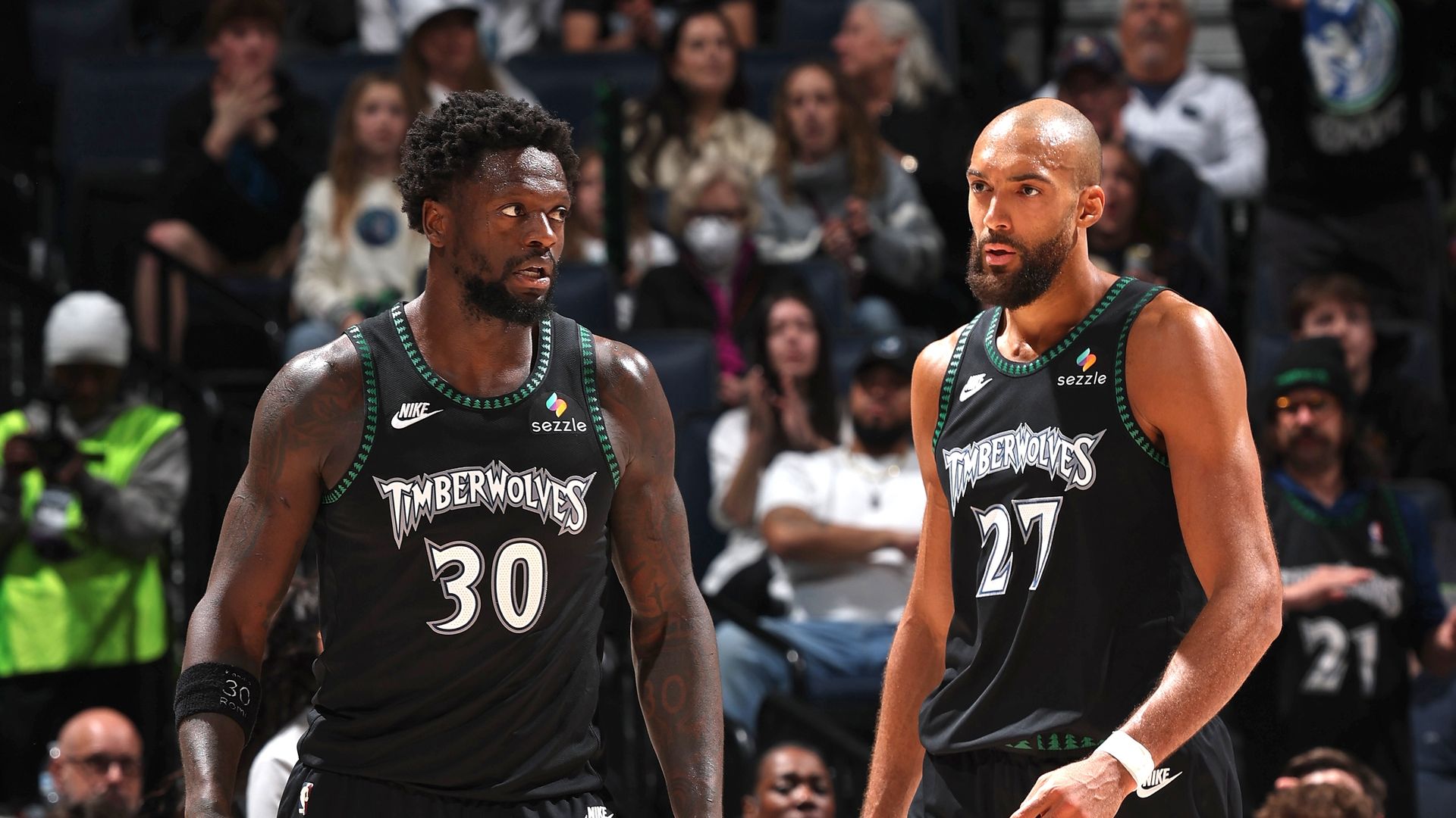 Two Timberwolves players on a basketball court wearing black jerseys with green trim, numbers 30 and 27. Crowded stands behind them; left player in green sneakers, right in tan shoes.