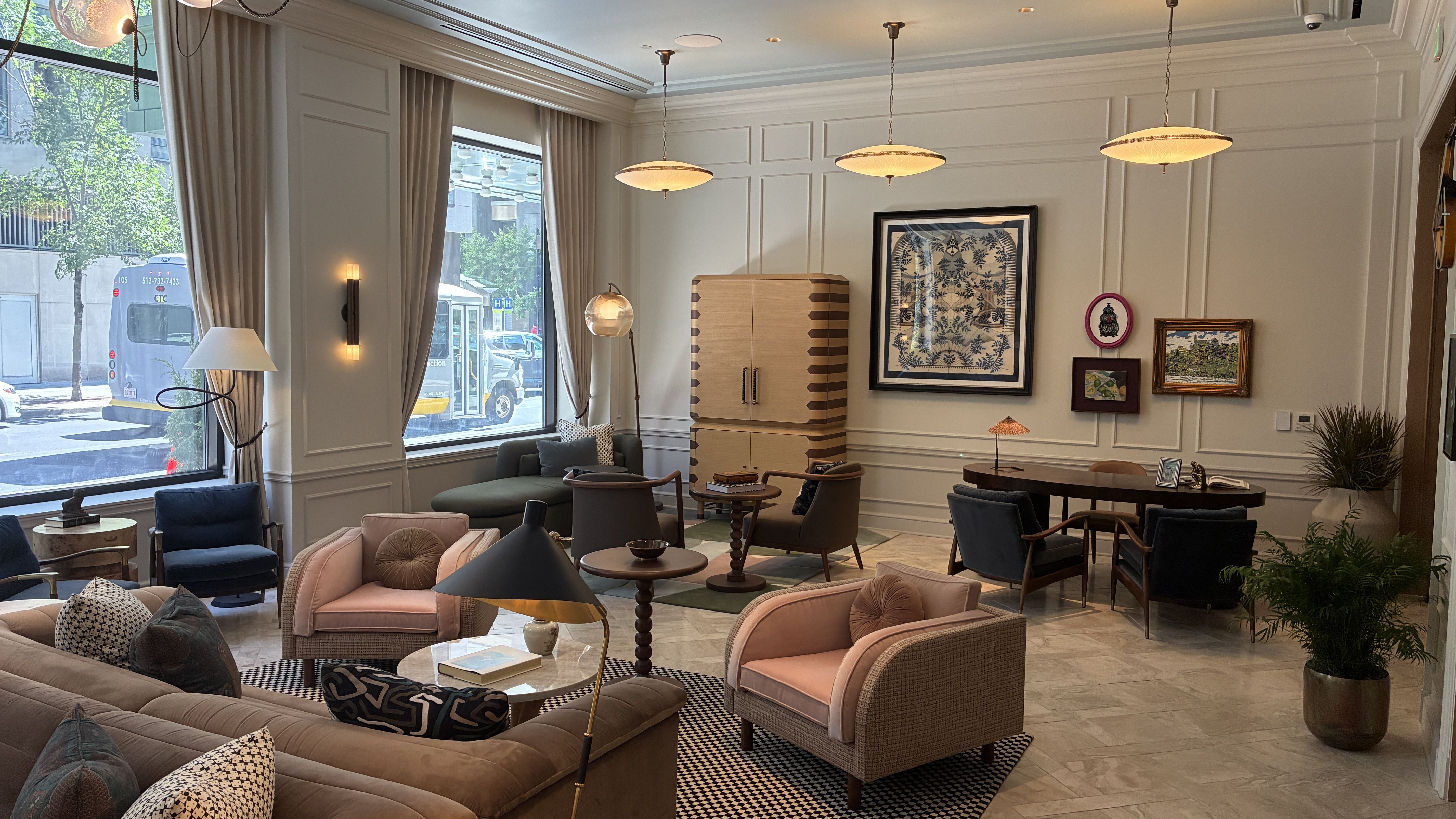 Bright hotel lobby with large windows and cream paneled walls. A striped wooden cabinet, framed blue botanical art, and pastel pink seating around round tables under warm pendant lights.