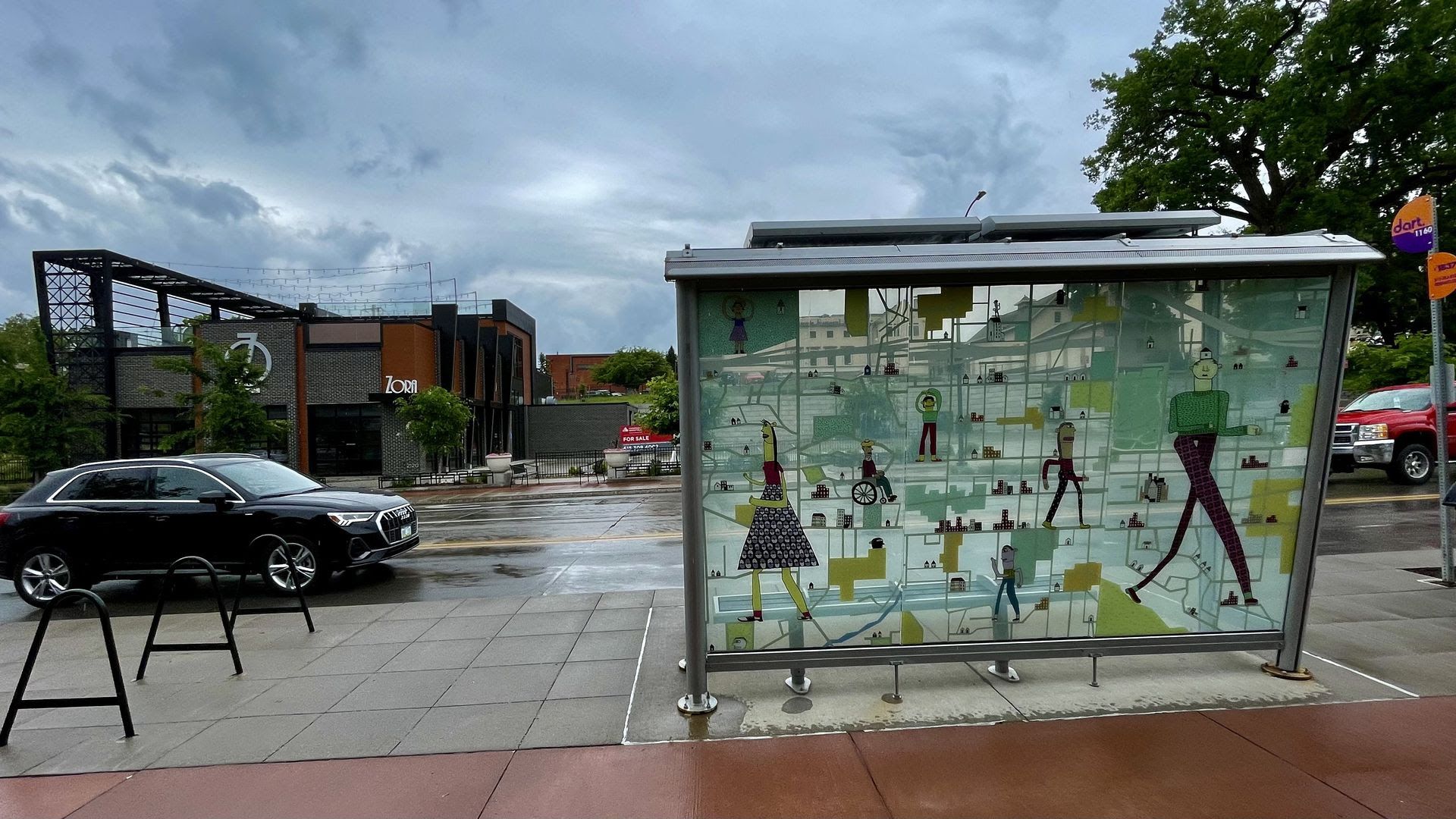 Bus stop shelter with colorful cartoon artwork of people in various poses over a light city map. Wet street and sidewalk, black and red vehicles, modern brick buildings, and cloudy sky.