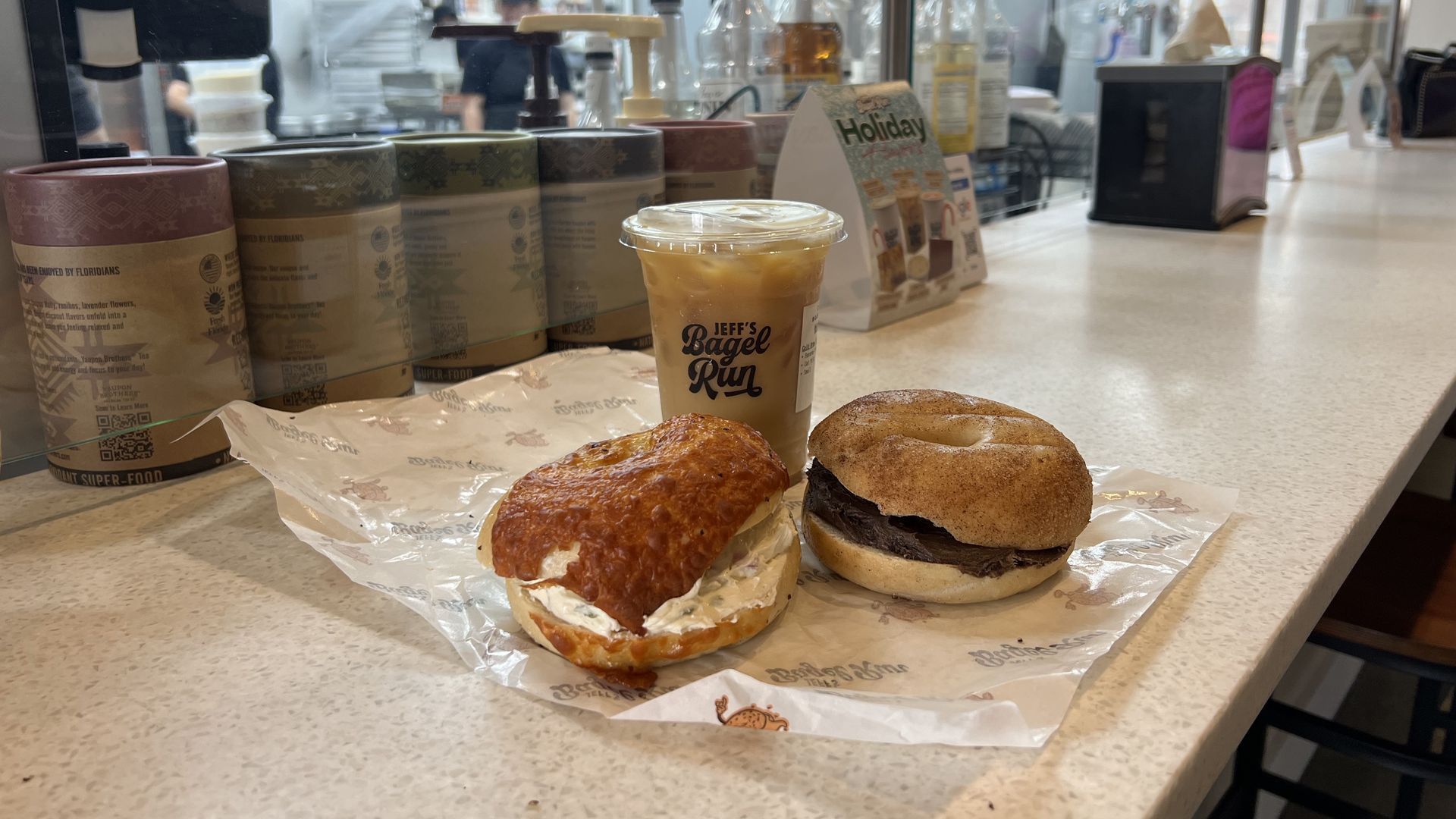Two bagels with cream cheese on parchment—left sesame, right cinnamon-raisin—beside an iced coffee cup labeled "Jeff's Bagel Run." Pastel canisters and a "Holiday" promo are visible in the background.